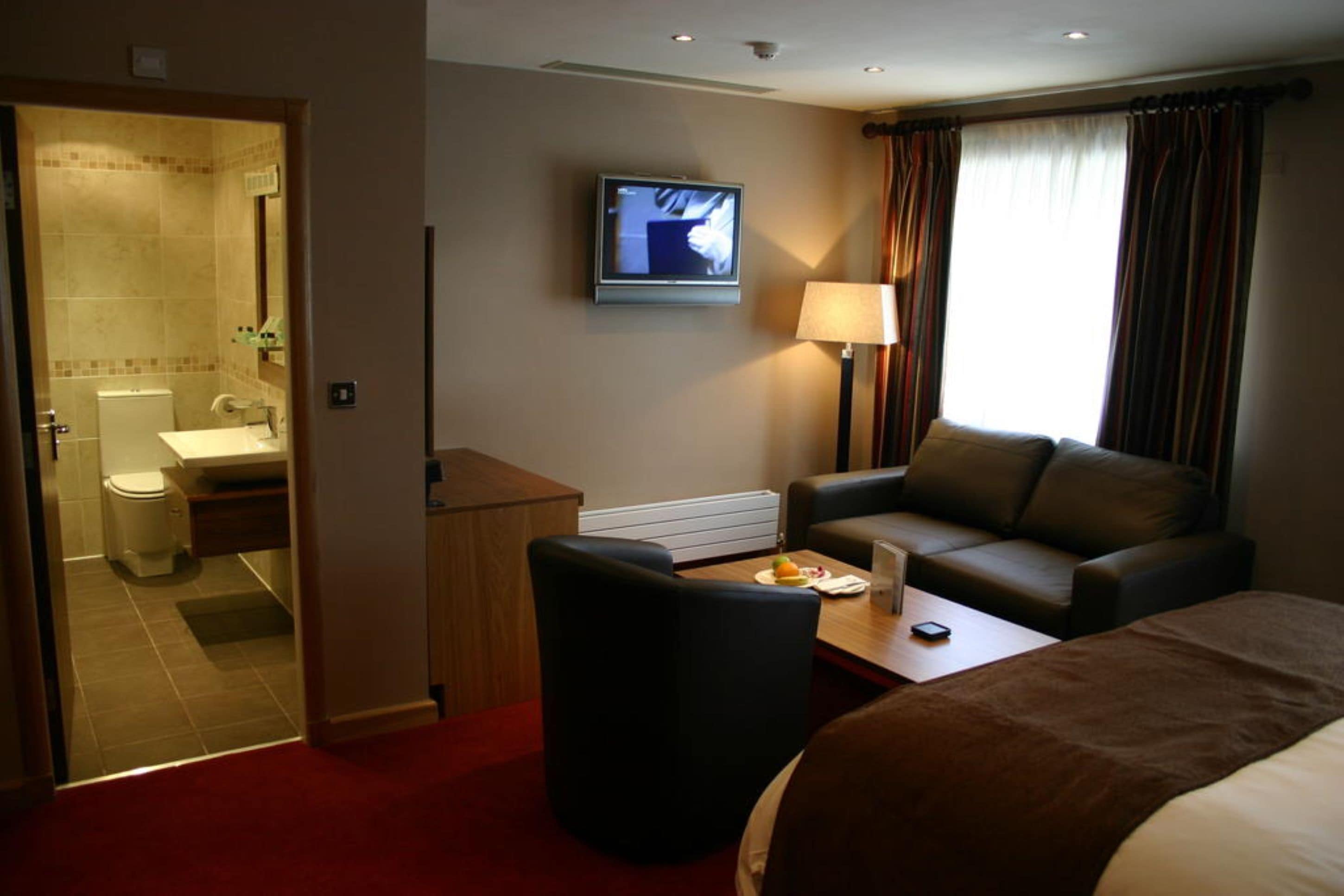 A hotel room with a bed, a sofa, a coffee table, a TV, a bathroom with a toilet and sink, and a window with curtains.