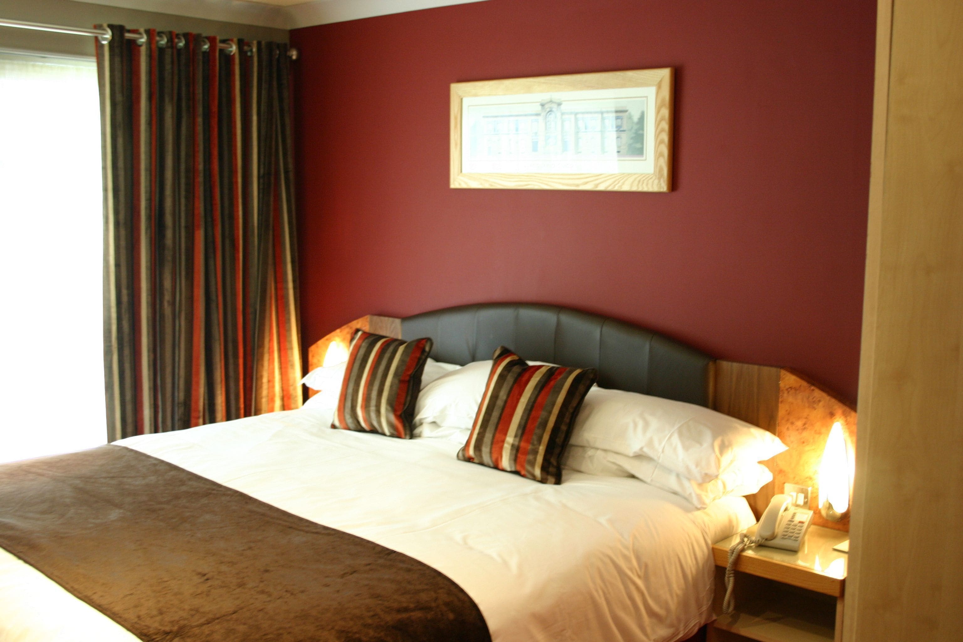 A hotel room with a large bed, striped pillows, a headboard, a bedside table with a lamp, and a framed picture on the wall.