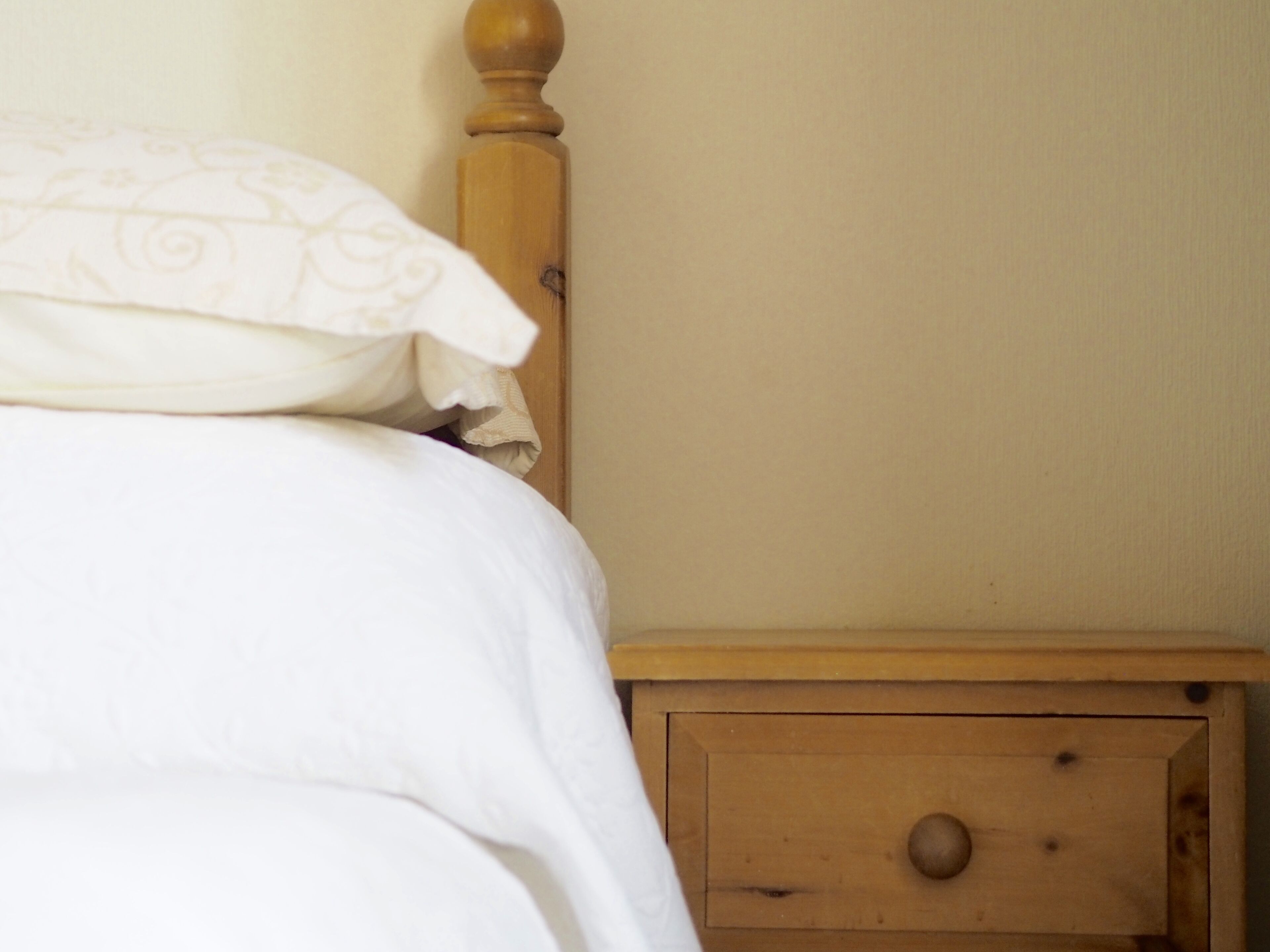 Una cama con ropa de cama blanca y una mesita de noche de madera con un cajón.