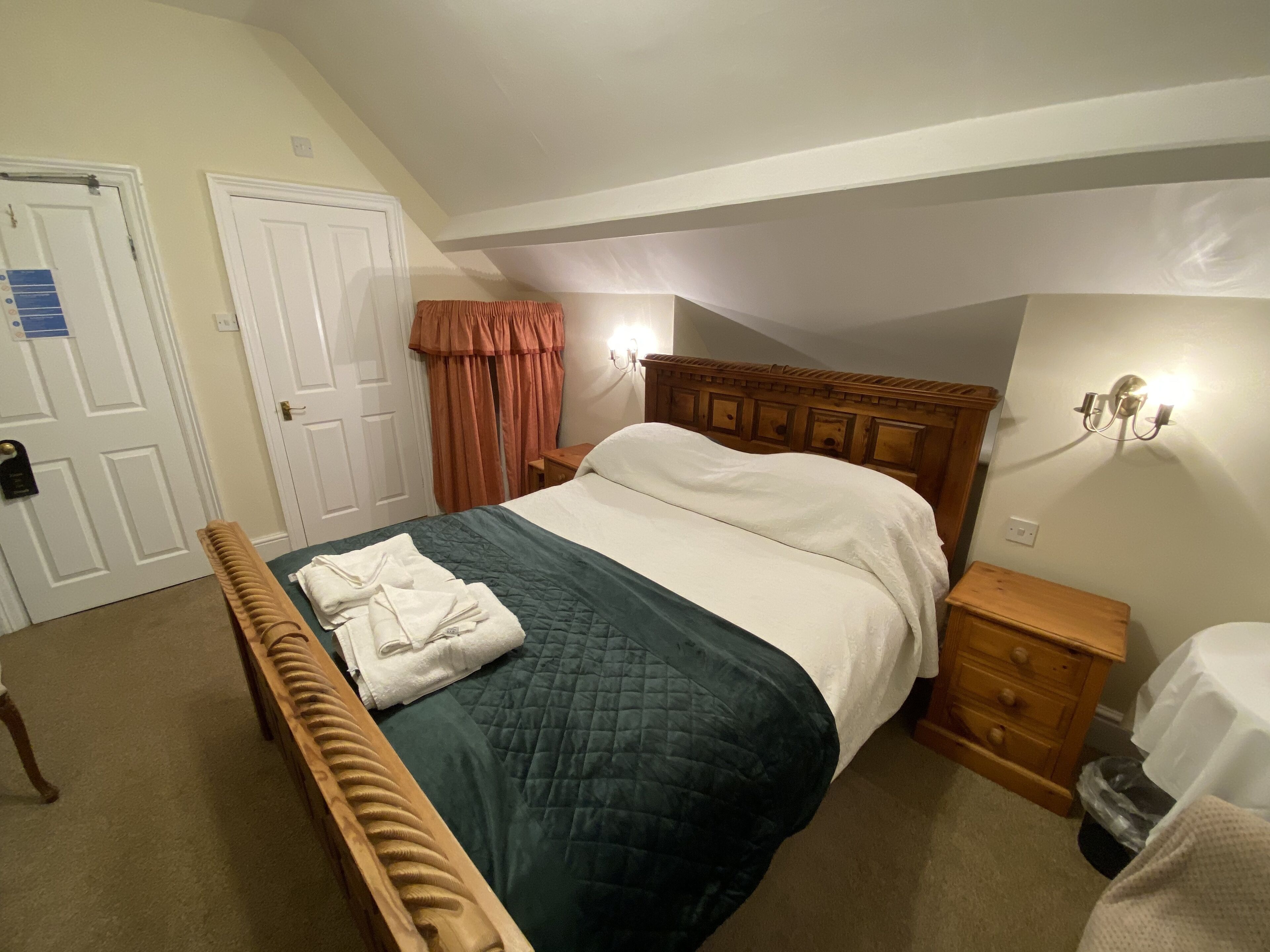 Un dormitorio con cama, cabecero de madera, mesita de noche, banco, luz empotrada en la pared y una puerta.