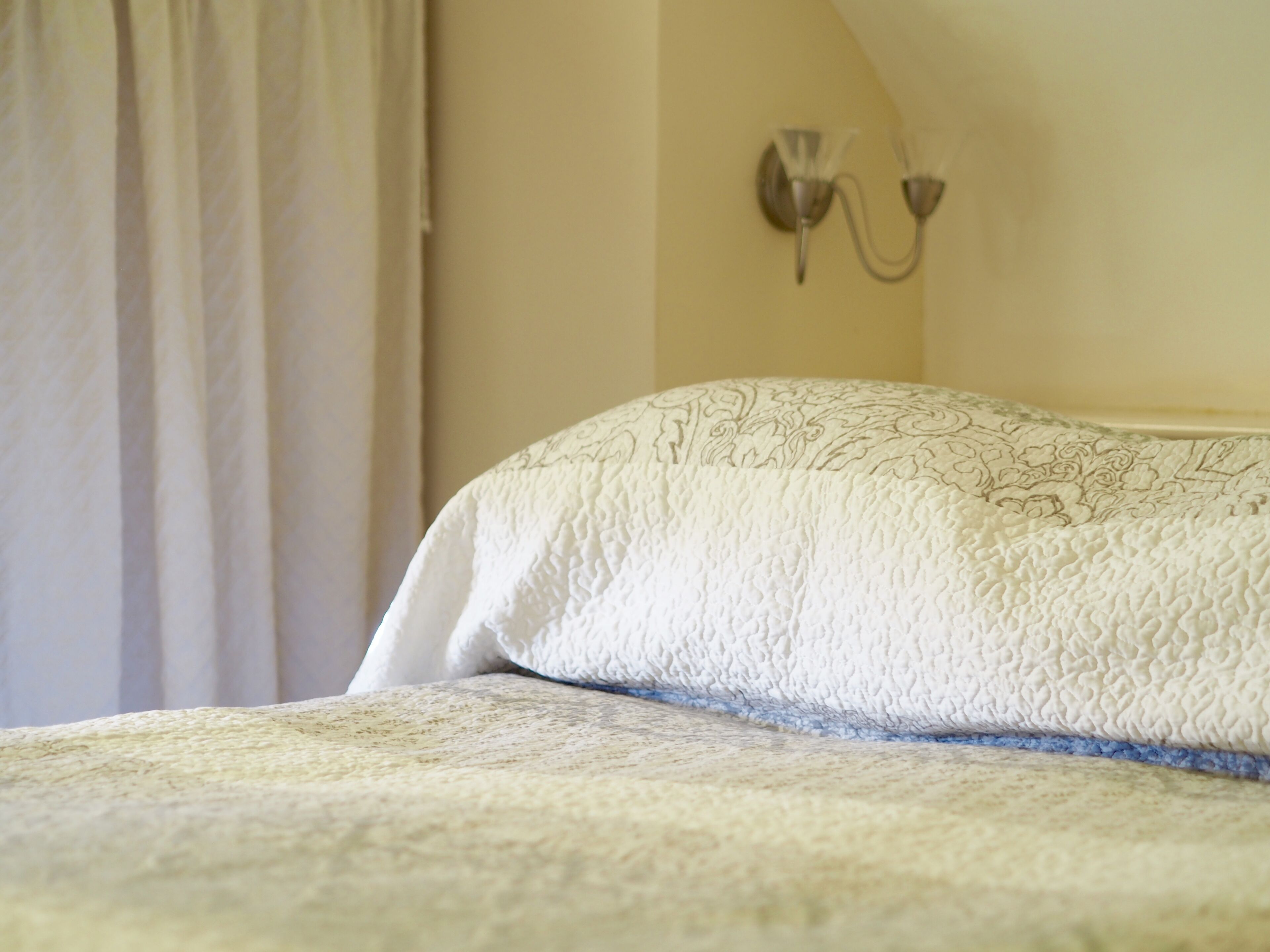Una cama con un edredón blanco texturizado y una almohada estampada, un aplique de pared y una cortina.