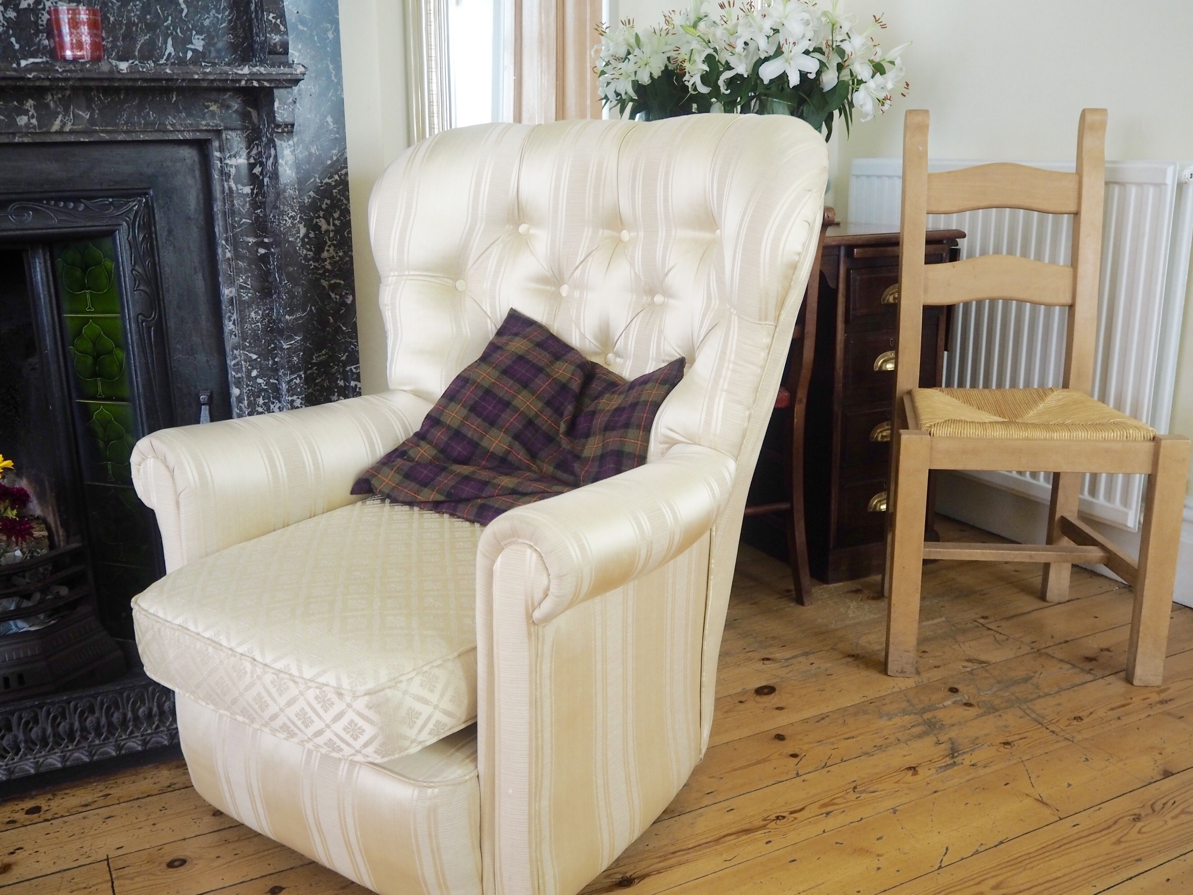 A white leather armchair with a plaid pillow, a wooden chair, a vase of white flowers, and a fireplace with decorative glass panels.