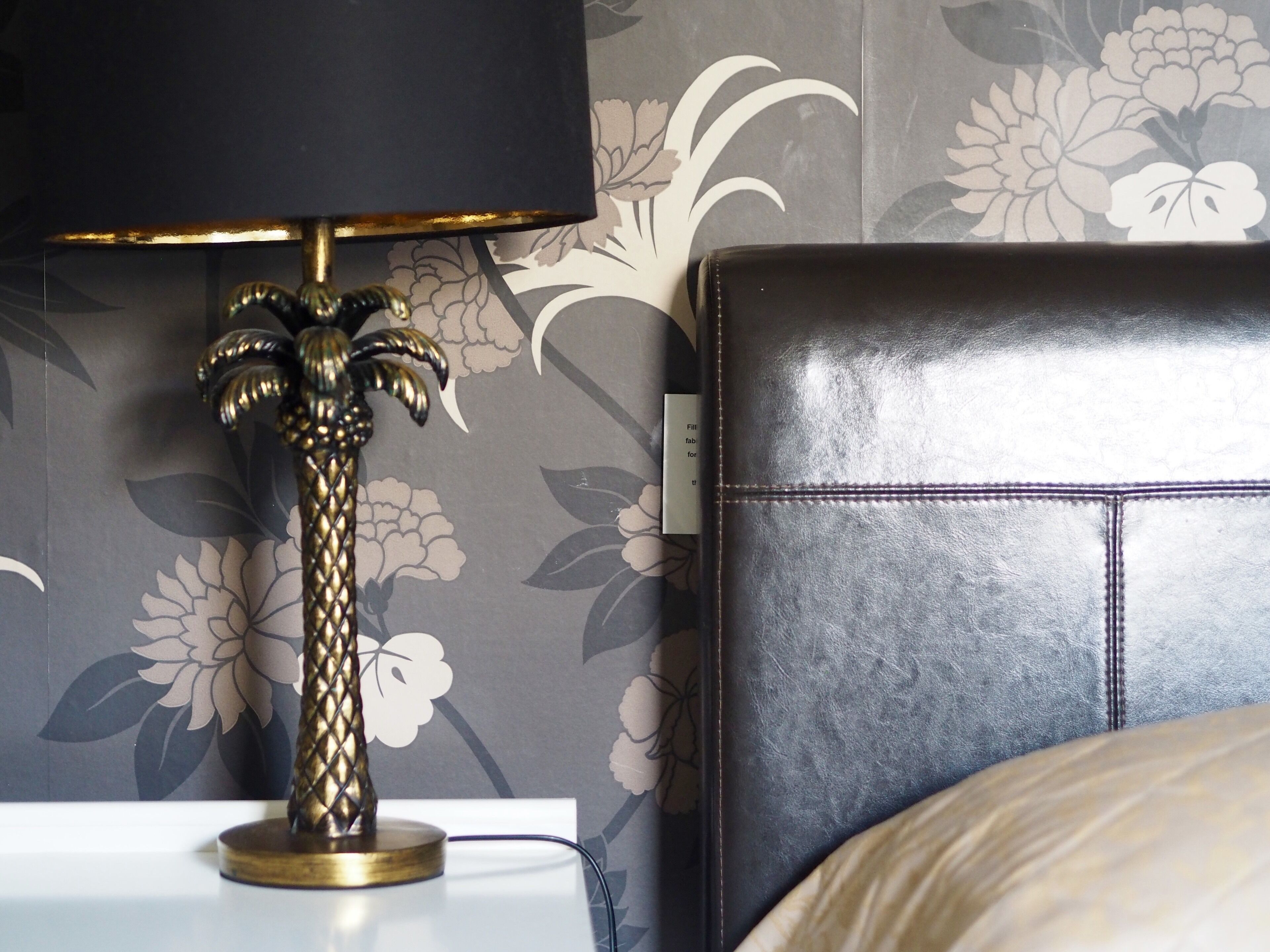 Una cama con cabecera negra, una lámpara decorativa con diseño de palmera y un estampado floral en el papel tapiz.