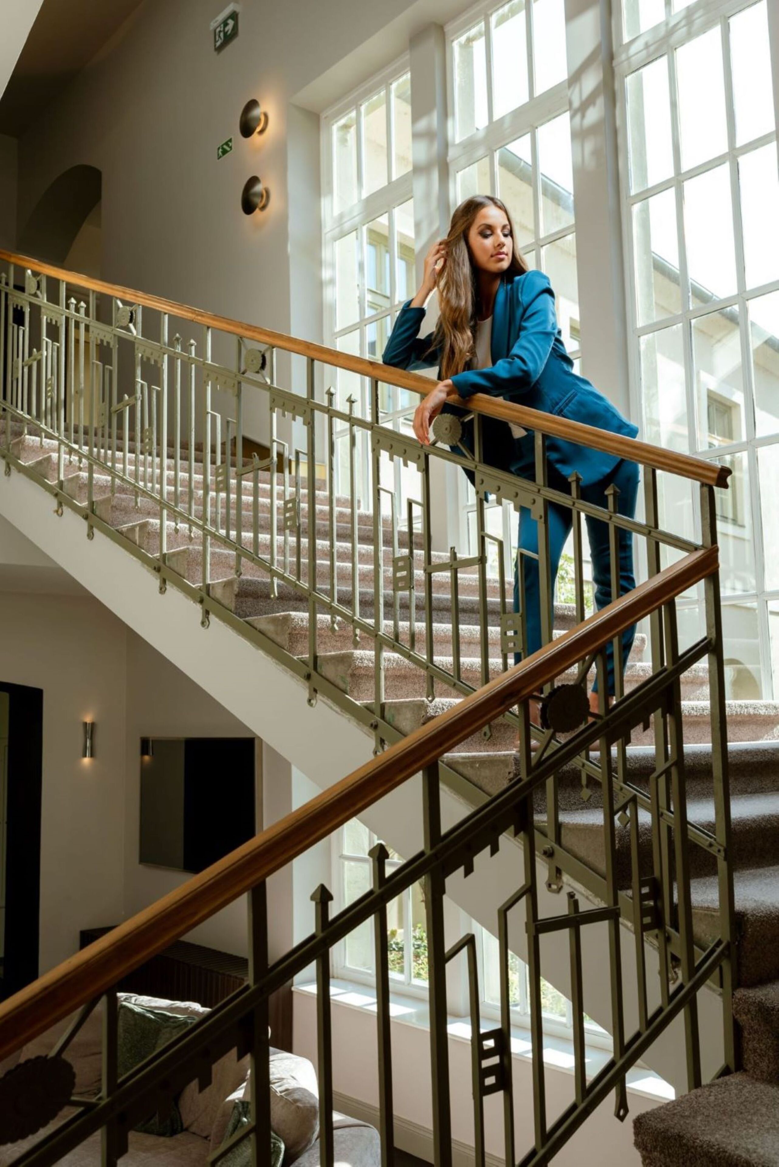 A woman standing on a staircase with a glass railing, a wooden handrail, and a view of a bright interior space with large windows.