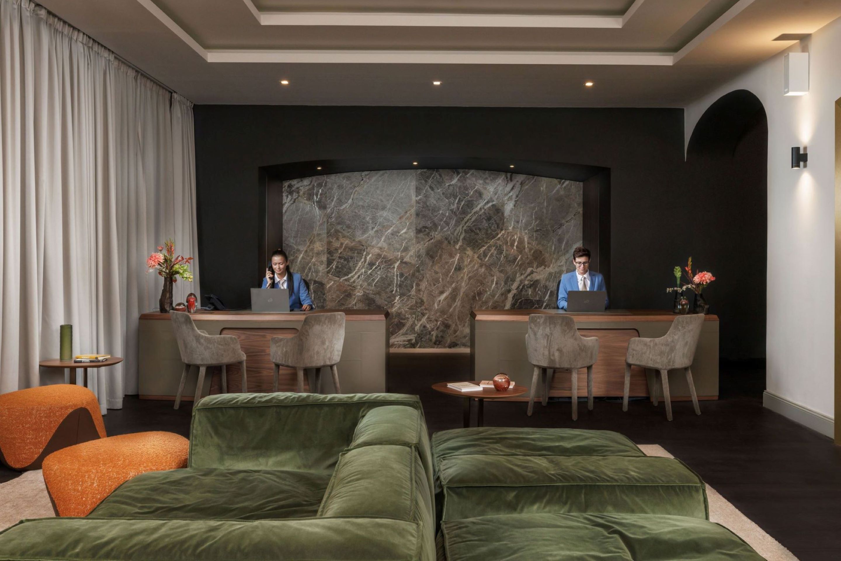A modern hotel lobby with a marble reception desk, two receptionists, a green sofa, and a small table with a vase.