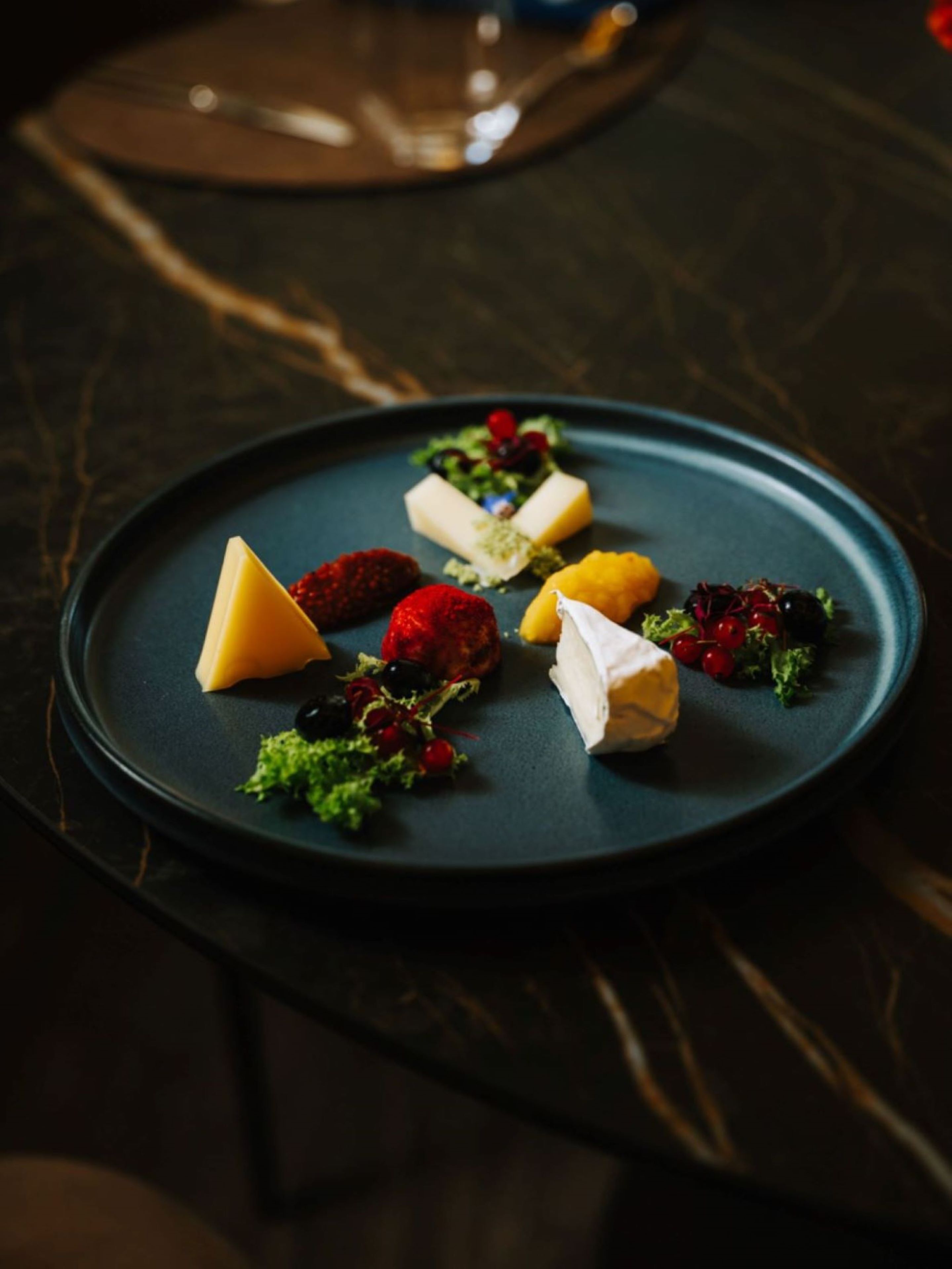 Una mesa de mármol oscuro con un plato azul que contiene diversos quesos y frutas.