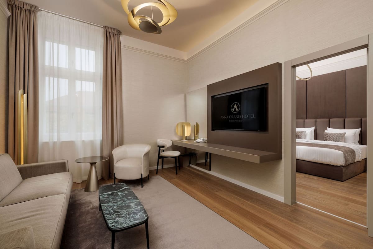 A modern hotel room with a sofa, a small table, a TV mounted on the wall, and a bed with a headboard.