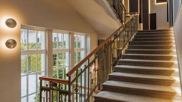A modern staircase with a wooden handrail and metal railings, leading to a landing with large windows overlooking greenery.