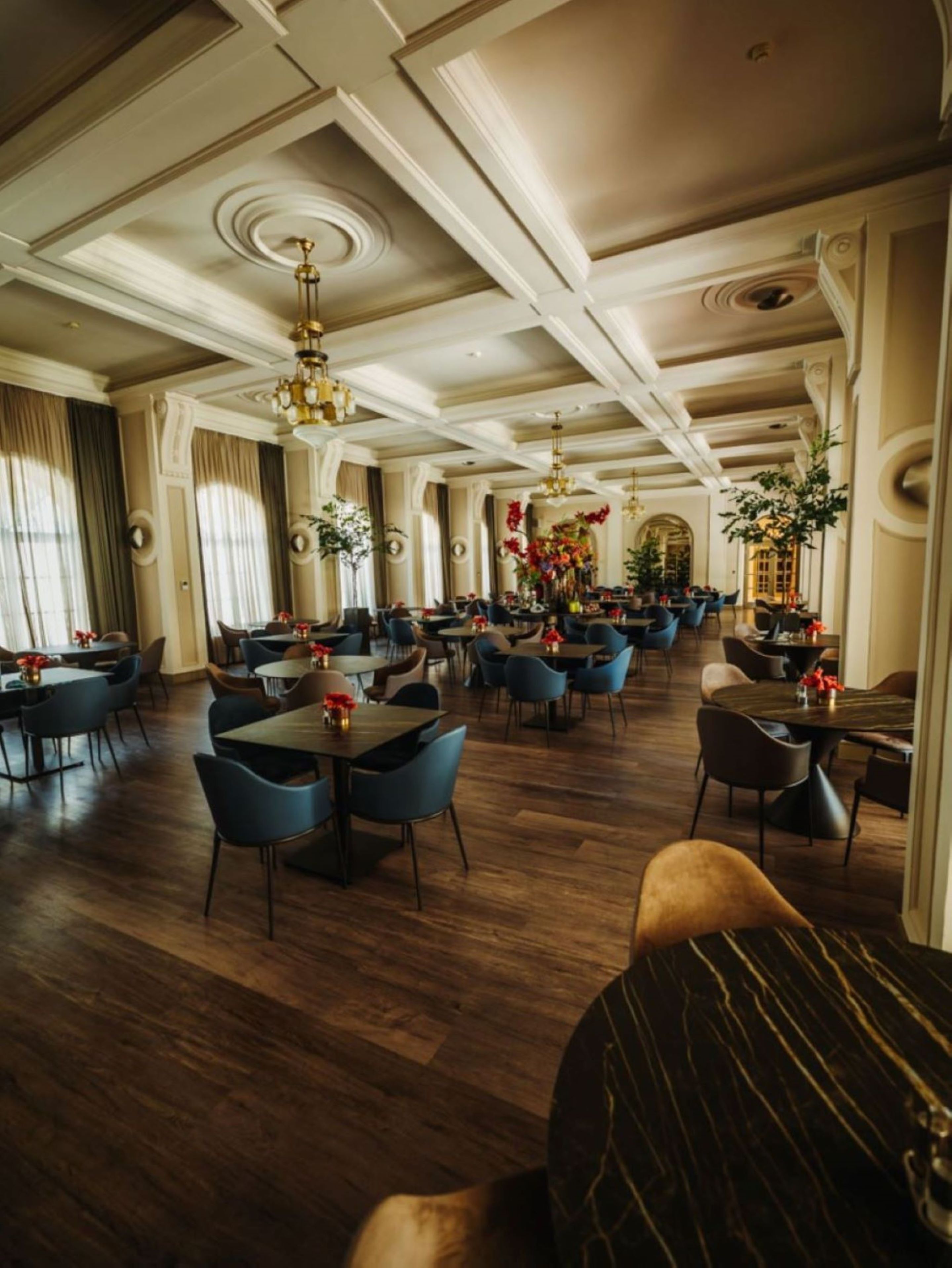 A spacious hotel dining area with multiple tables and chairs, elegant chandeliers, and decorative plants.