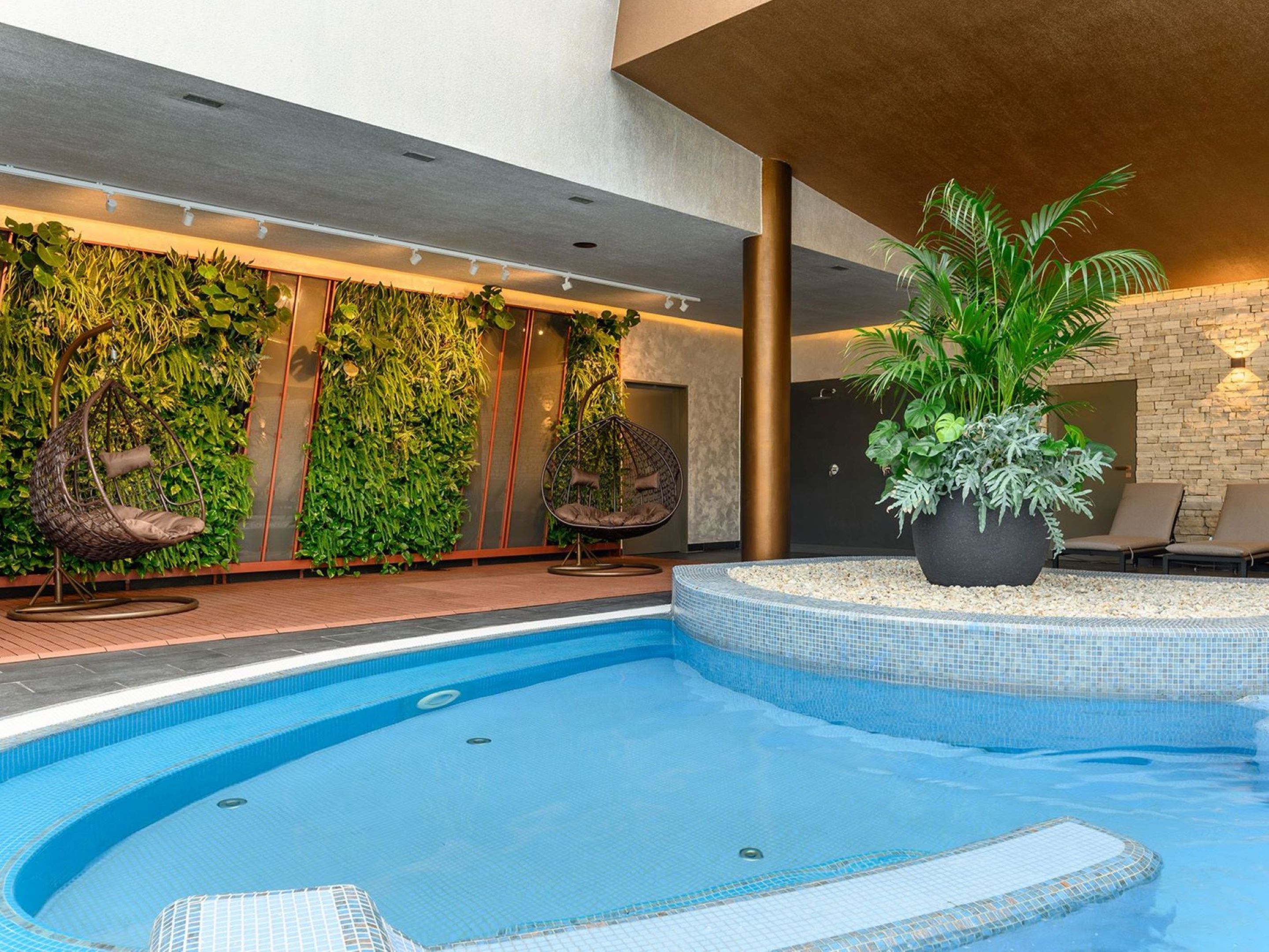 Una piscina interior con borde de azulejos, rodeada por una terraza de madera. Hay dos sillones colgantes y una planta grande en maceta cerca de la piscina.