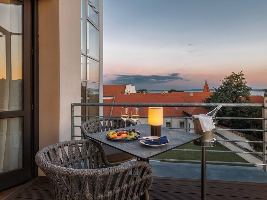 A balcony with a table set for two, a chair, a lamp, and a view of buildings and a sunset.