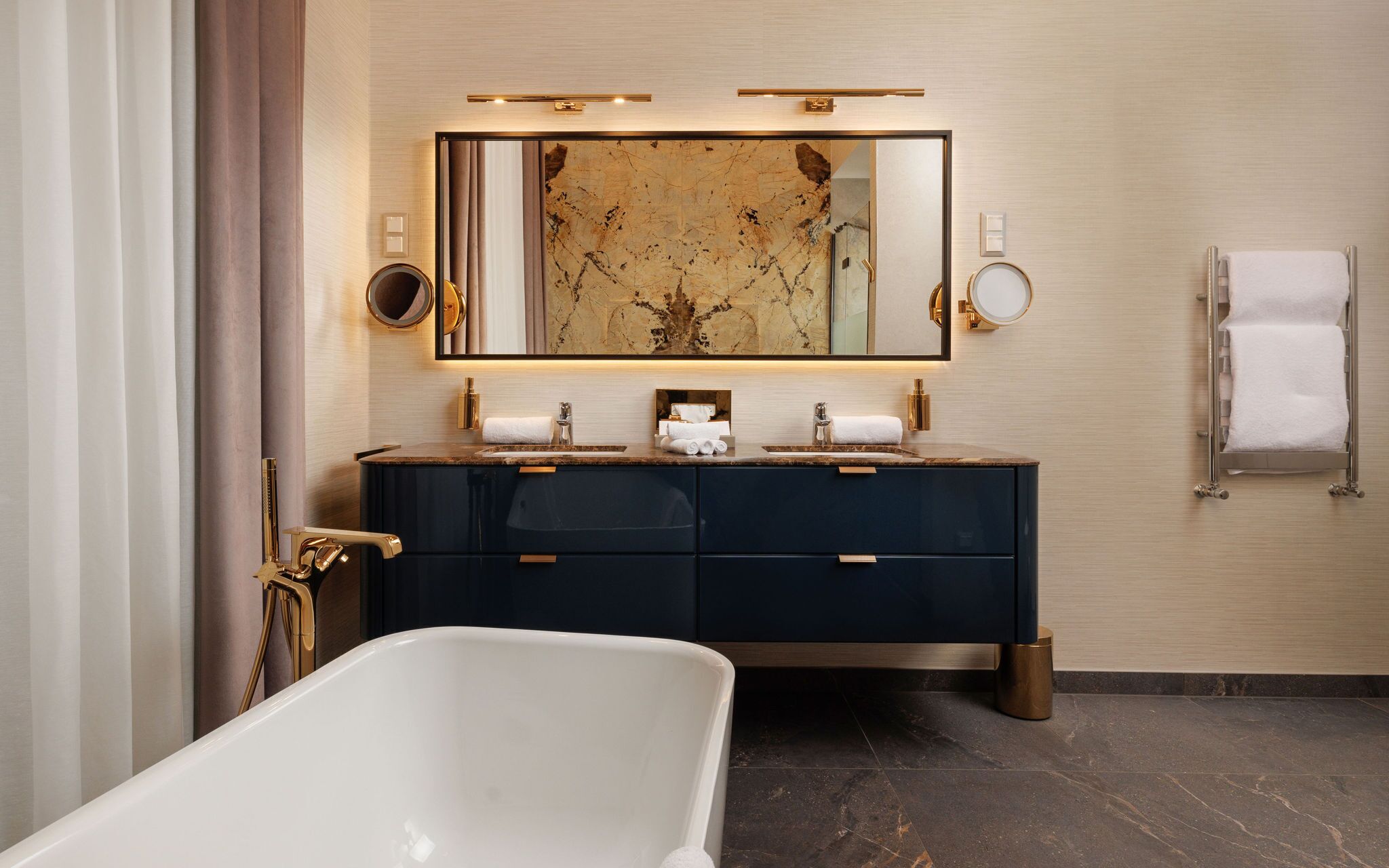 A modern bathroom with a freestanding bathtub, dark blue vanity with gold fixtures, and a large mirror above the sink.