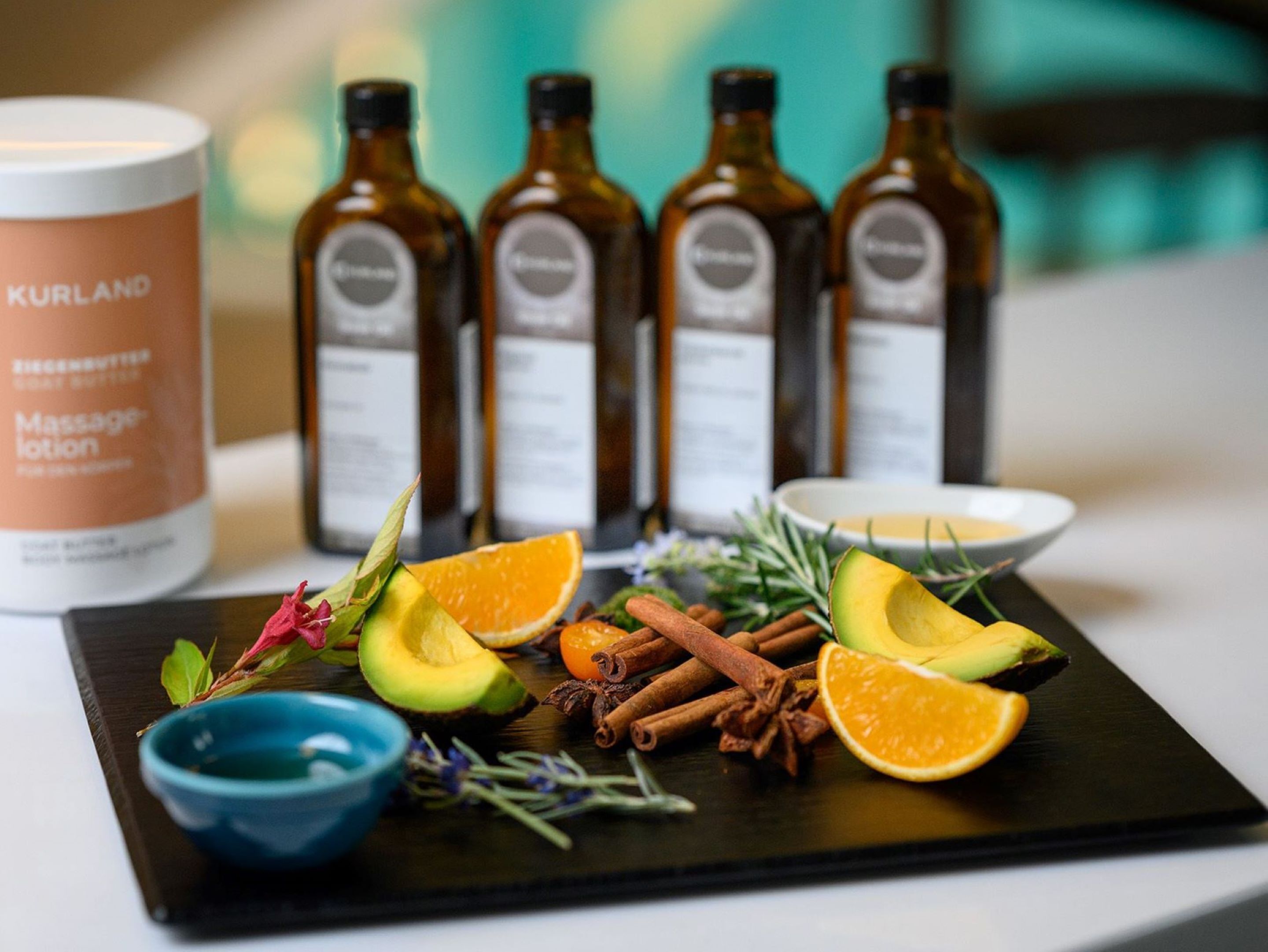 Un plateau contenant des tranches d’avocat, des oranges, des bâtons de cannelle et des herbes, ainsi que des bouteilles de lotion de massage Kurland et un bol bleu.