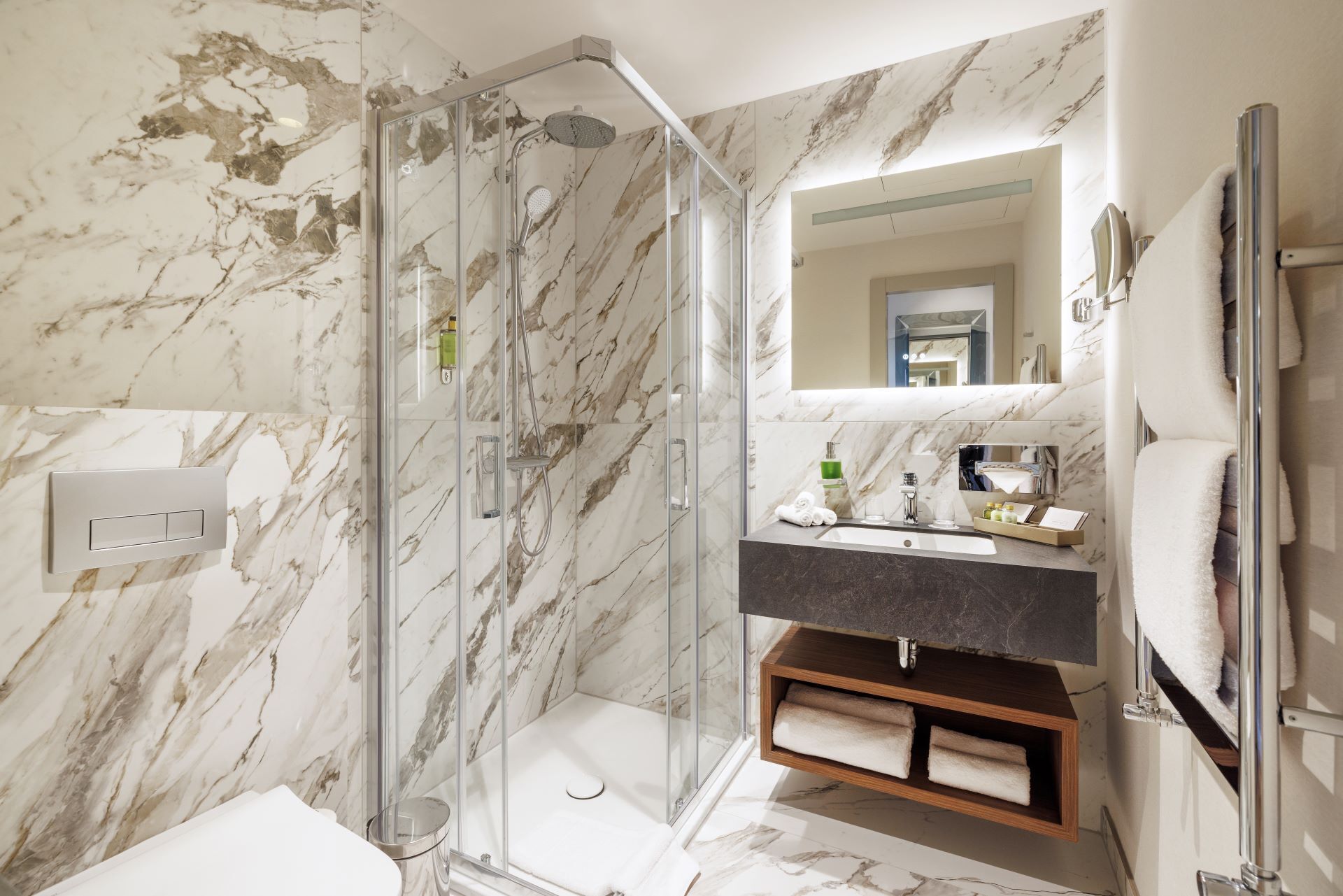Ein modernes Badezimmer mit einer Glaskabine zur Dusche, Marmorwänden, einem Waschtisch mit Spiegel und einem Holzregal.