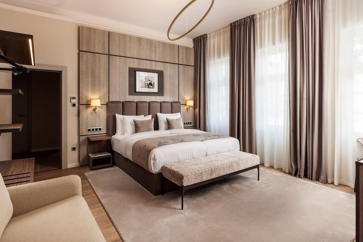 A modern hotel room with a large bed, a bedside table, a nightstand, a sofa, and a large window with curtains.
