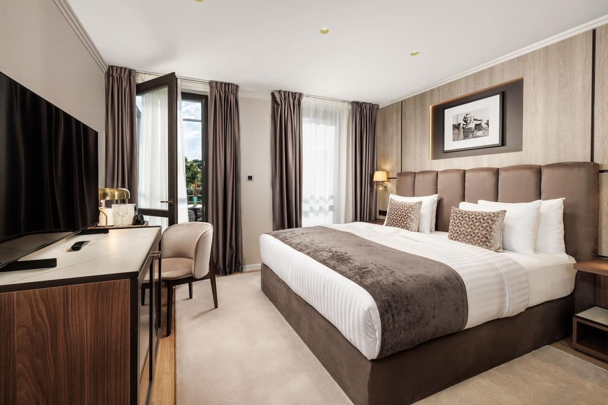 A hotel room with a bed, bedside table, chair, desk, and a framed picture on the wall.