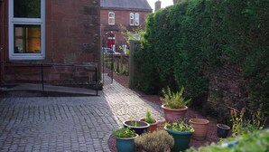 A cobblestone pathway leading to a building with a brick facade and a large window. There are potted plants along the pathway and a hedge to the right. - The Old Rectory (Annan)