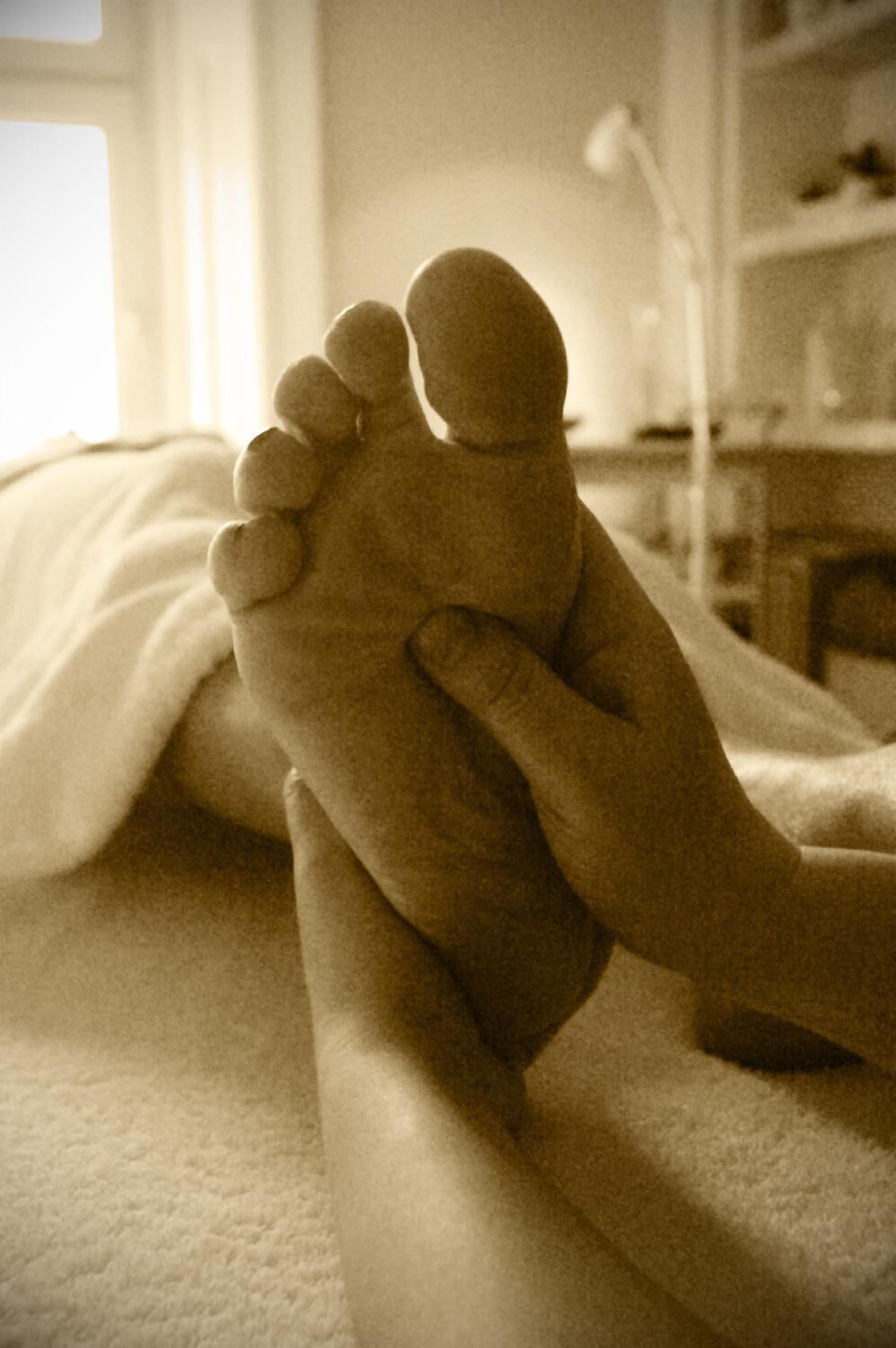 A foot resting on a bed with a blanket, in a room with a window and a desk with a lamp.