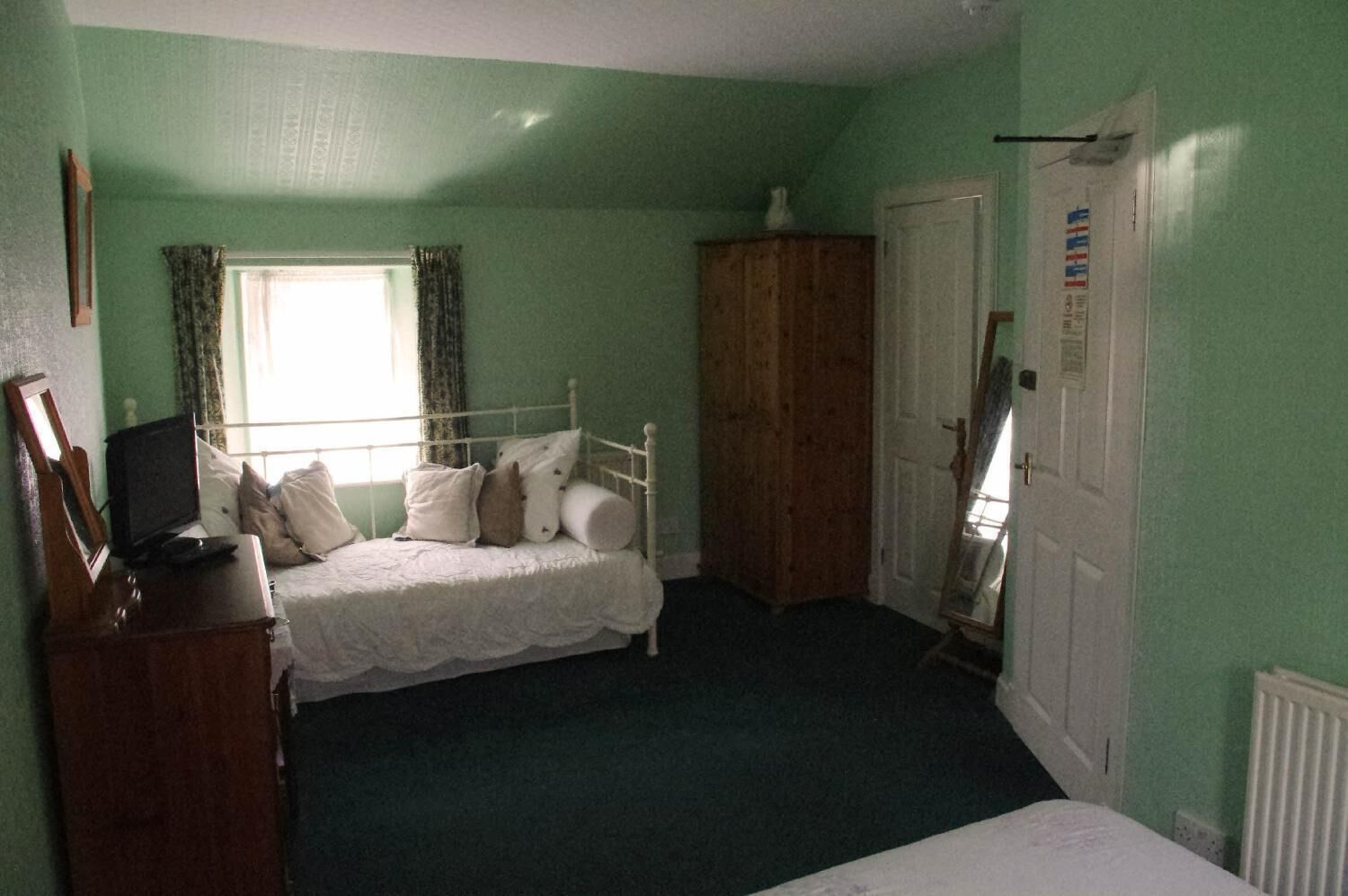 Une chambre avec un lit simple, une armoire en bois, un miroir et une fenêtre avec des rideaux.