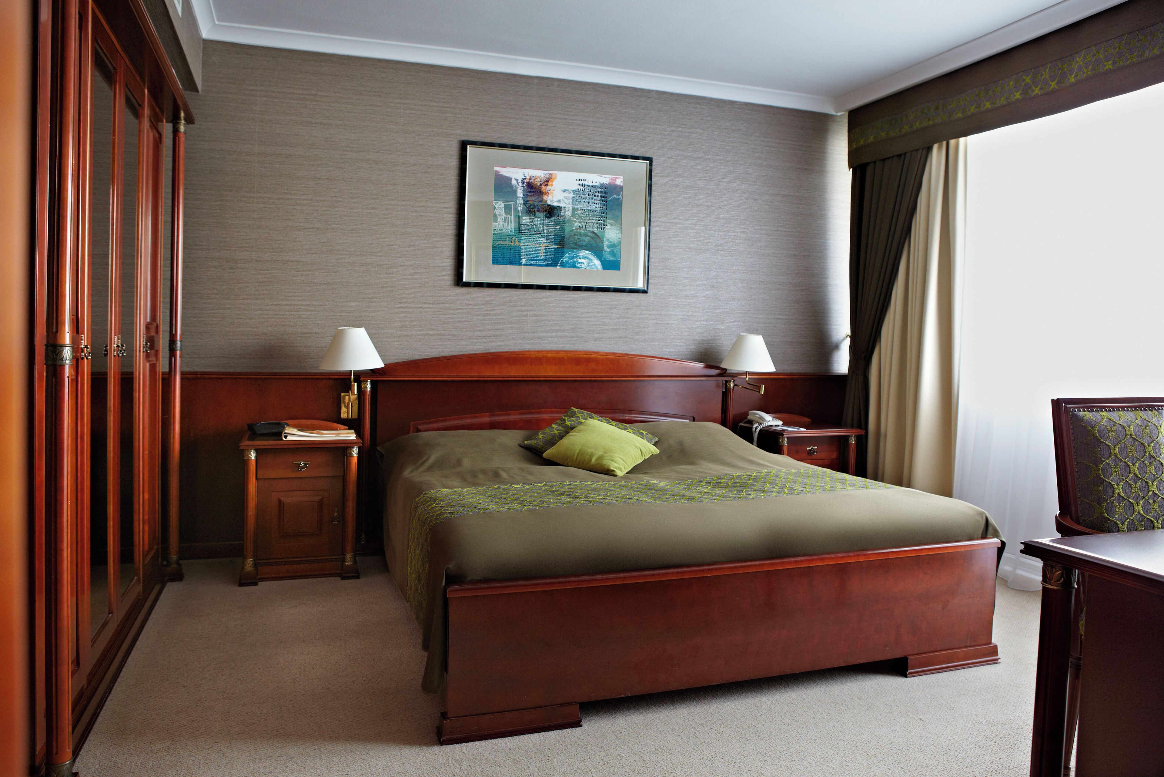 Une chambre d’hôtel avec un grand lit, une tête de lit en bois, deux tables de chevet avec des lampes, un tableau encadré au mur et une vue sur l’extérieur par une fenêtre avec des rideaux.