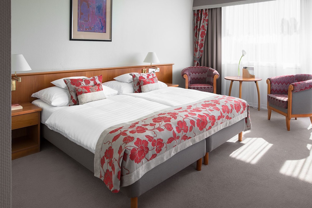 Une chambre d’hôtel avec un grand lit, deux chaises, une petite table et une fenêtre avec des rideaux.
