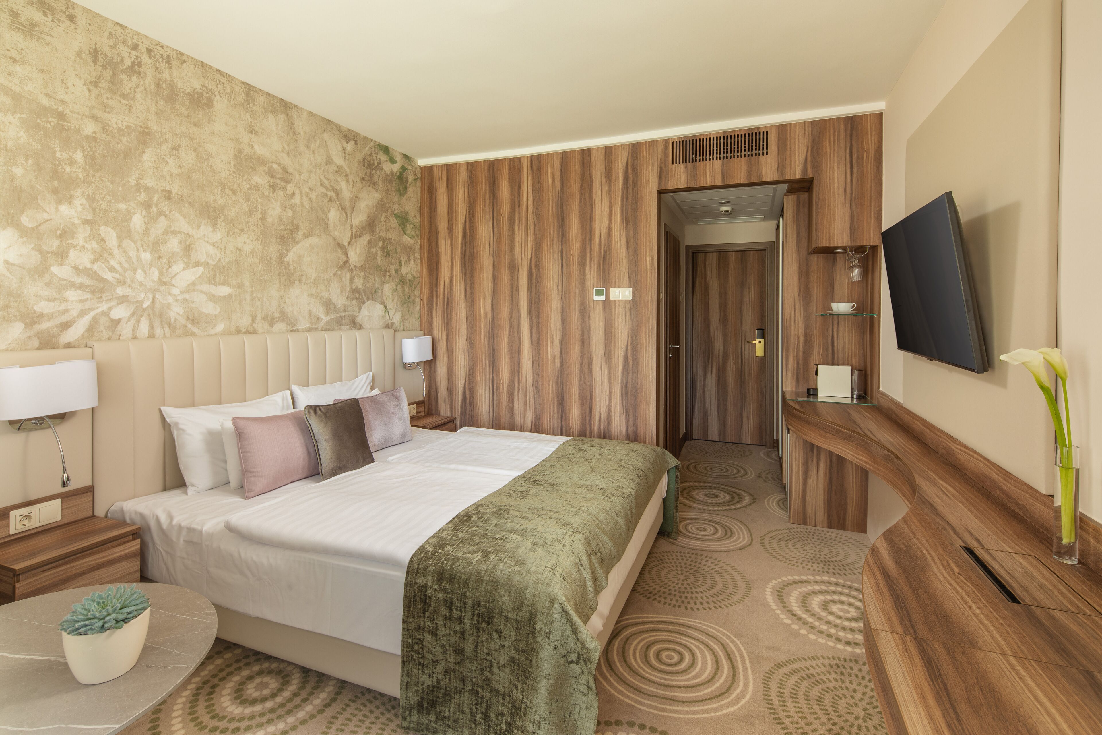Habitación de hotel con cama, mesita de noche, lámpara, televisión y una pequeña mesa con una planta.