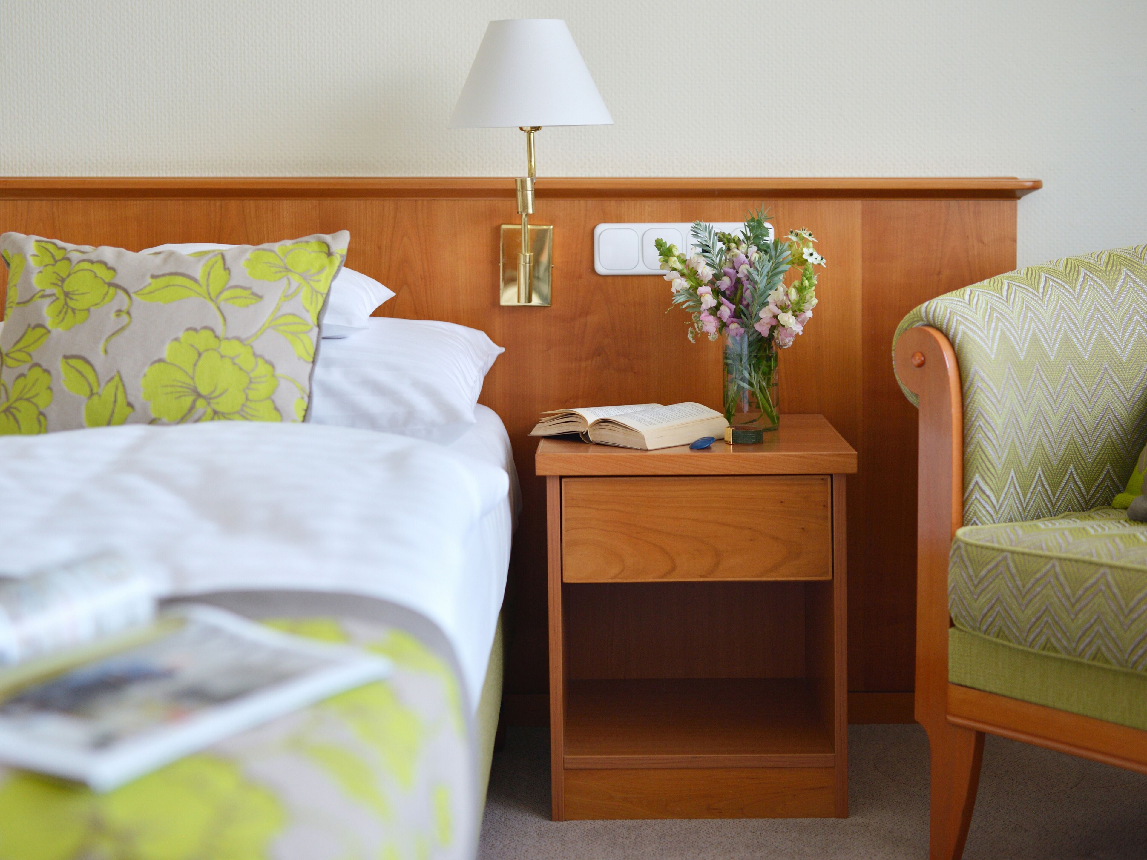 A hotel room with a bed, a nightstand with a lamp, a vase with flowers, and a chair.