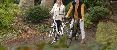 Deux personnes se promènent sur un chemin pavé, accompagnées de leurs vélos, et sont entourées de verdure et d’arbres.