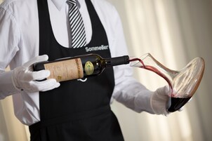 Una persona con camisa blanca y delantal negro sostiene una botella de vino y sirve en una copa.