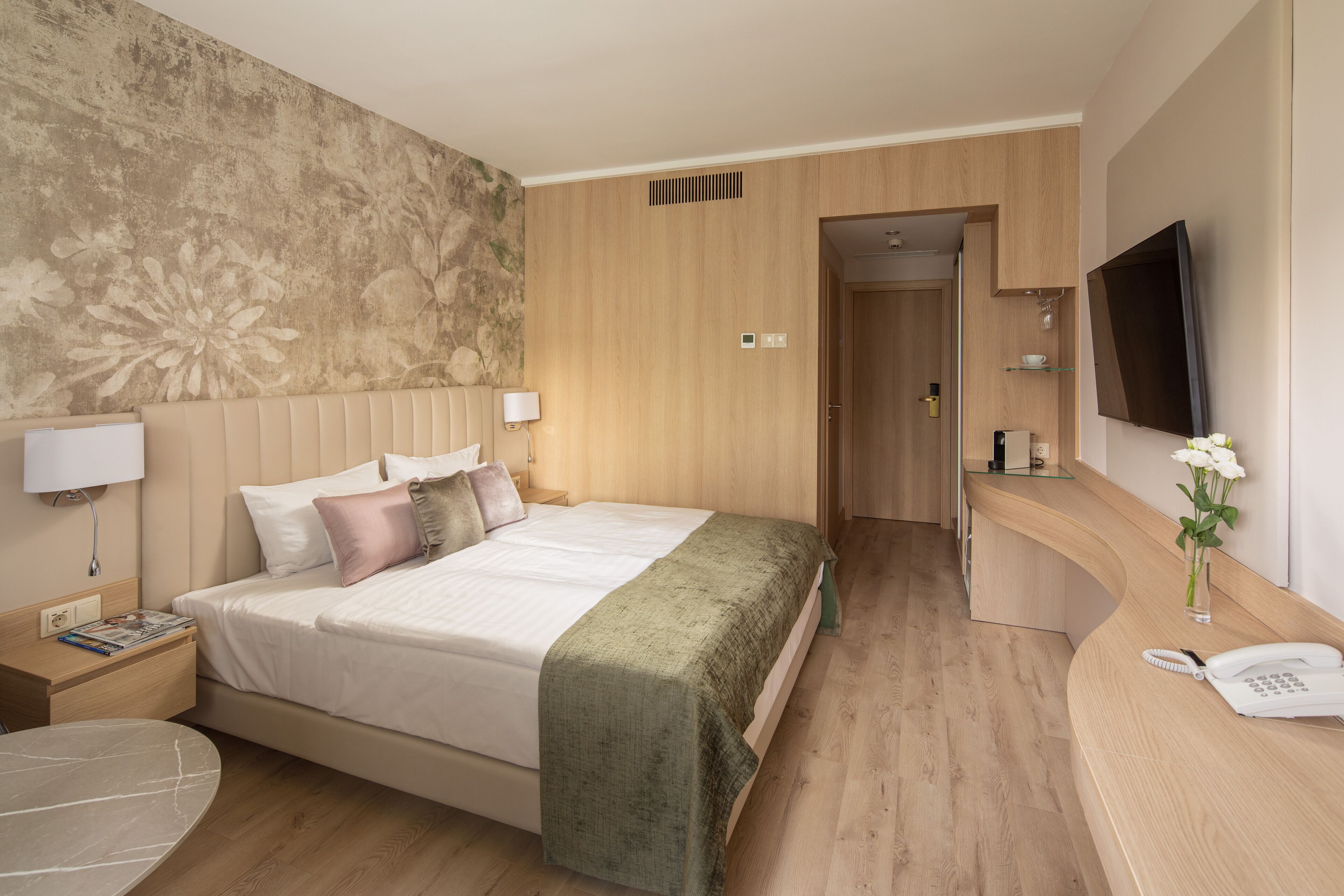 A hotel room with a large bed, bedside tables, a desk, a flat-screen TV, and a bathroom door.