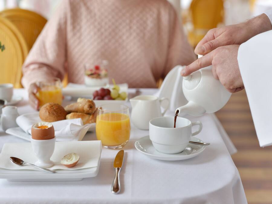 A breakfast setting with a table set for two, including plates, cups, a jug of orange juice, and a bowl of fruit.