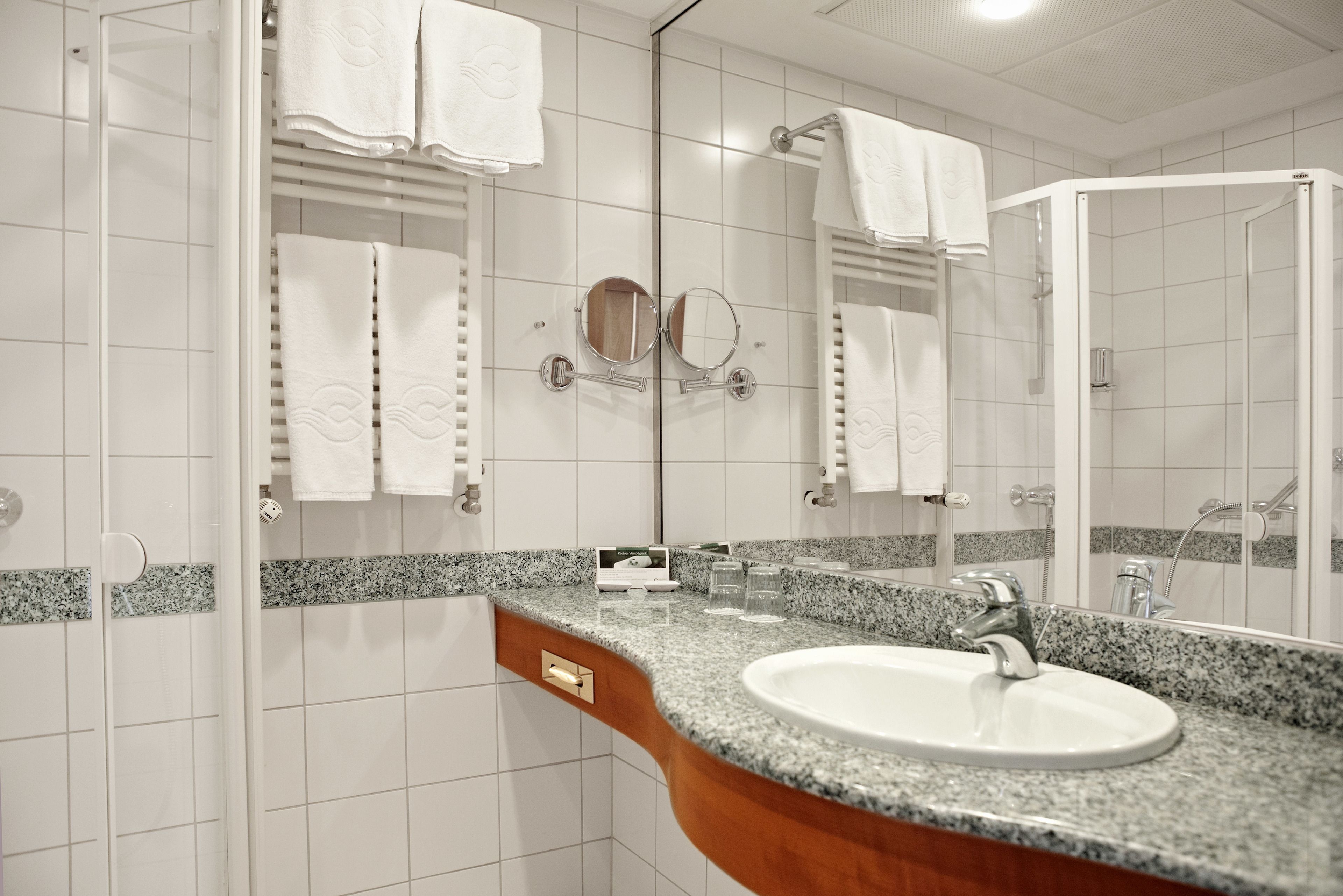 Un baño con lavamanos de encimera de granito, dos espejos y paredes con azulejos blancos.