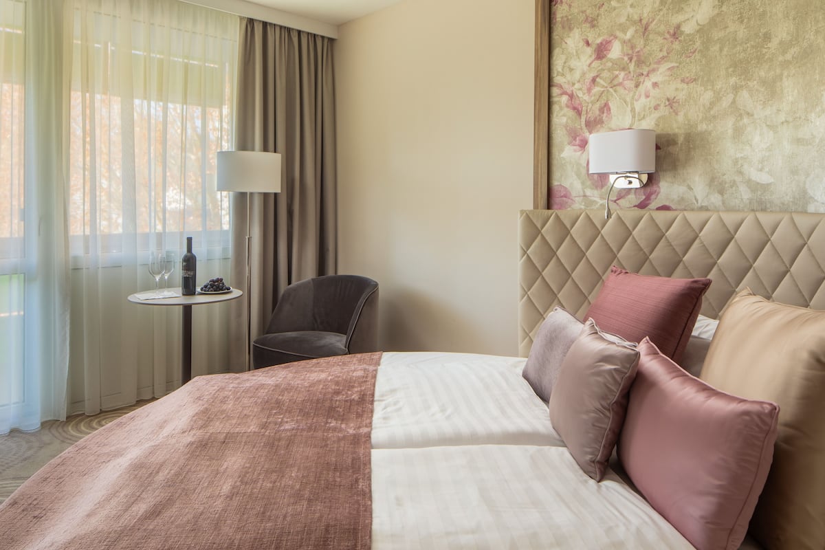 Une chambre d’hôtel avec un lit, une chaise, une petite table sur laquelle se trouvent une bouteille et des verres, et une lampe fixée au mur.