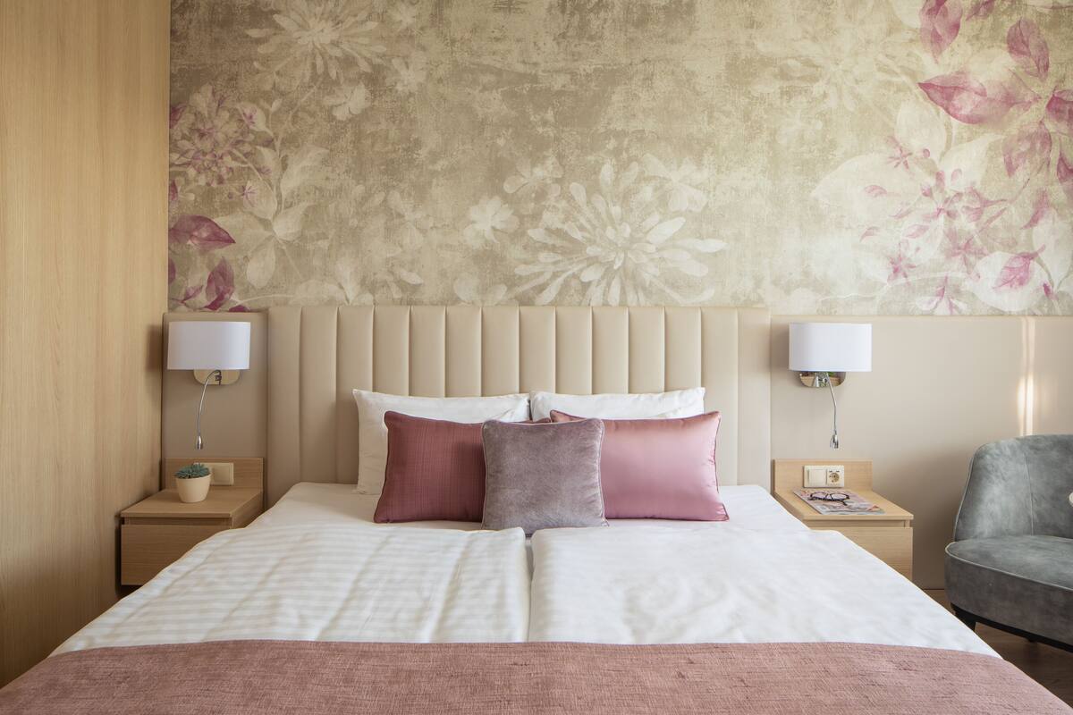 Un lit bien fait, avec une tête de lit à motifs floraux, deux tables de chevet avec des lampes et un fauteuil gris.