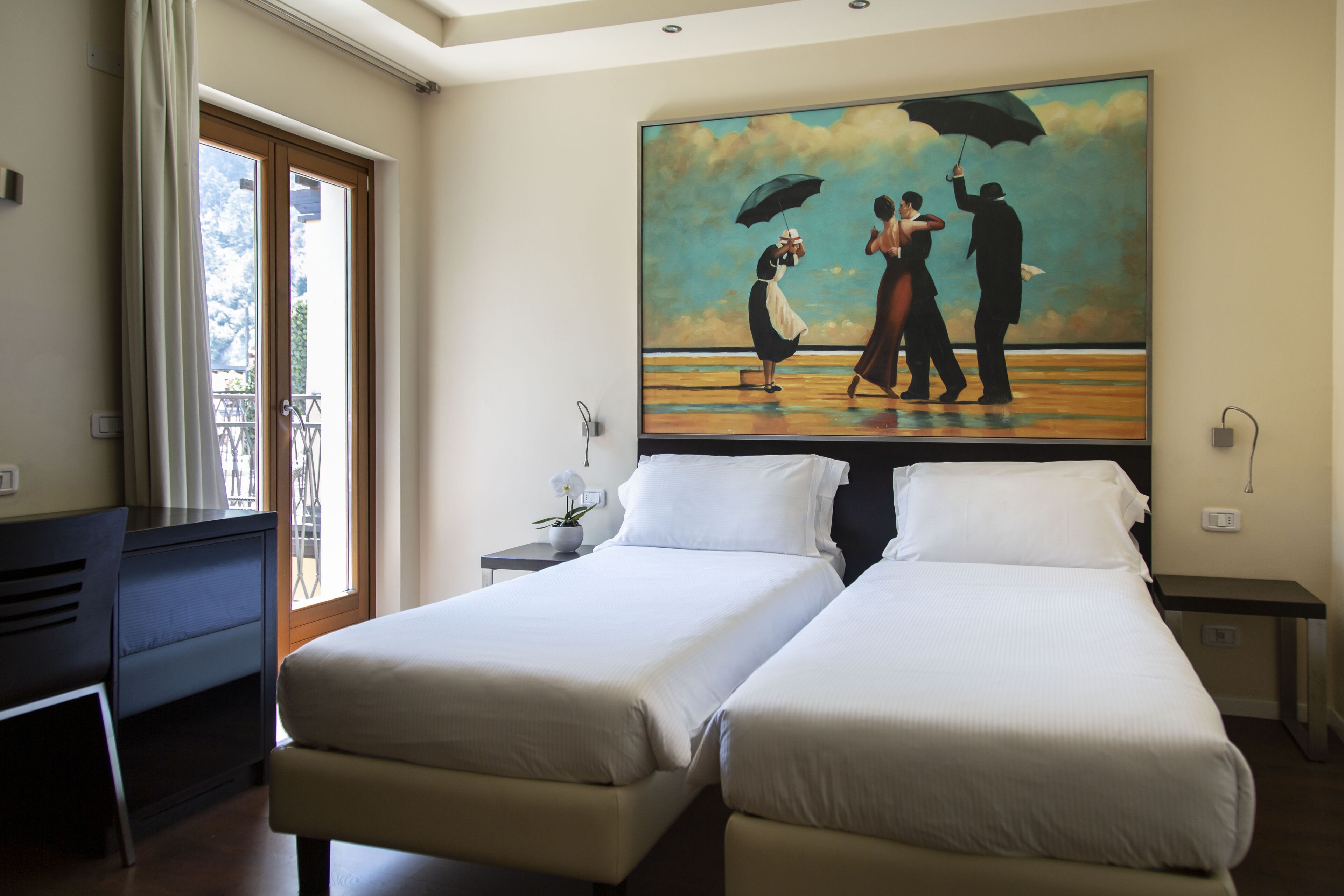 Hotelkamer met twee bedden, een bureau, een schilderij van een koppel op het strand en een balkon met uitzicht.