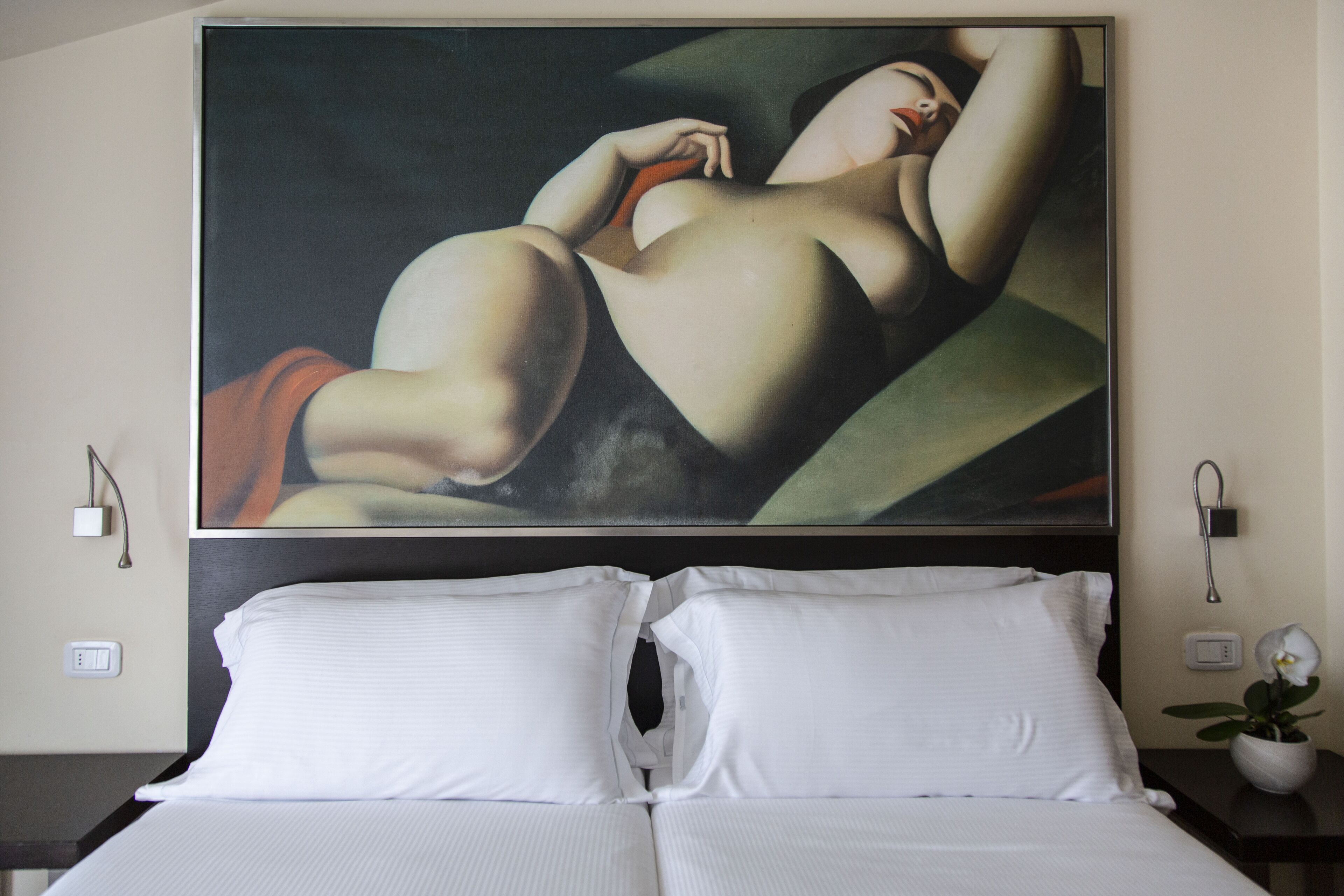 Une chambre d’hôtel avec un grand tableau au mur, un lit avec du linge de maison blanc, une table de chevet avec une plante en pot et une prise électrique au mur.