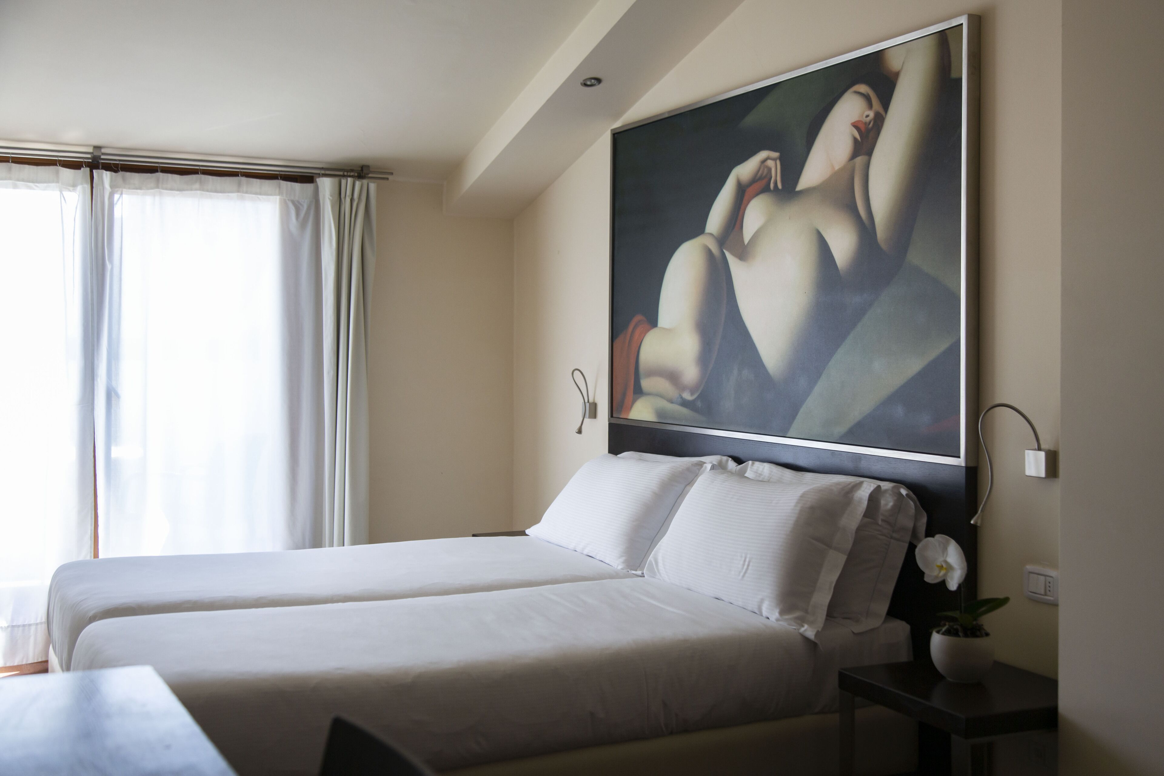 A hotel room with two beds, a large painting on the wall, a potted orchid on a nightstand, and a window with curtains.