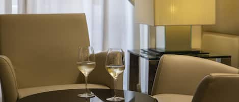 A hotel room with a round table, two chairs, and two glasses of white wine. There is a lamp on a side table and a window with curtains.