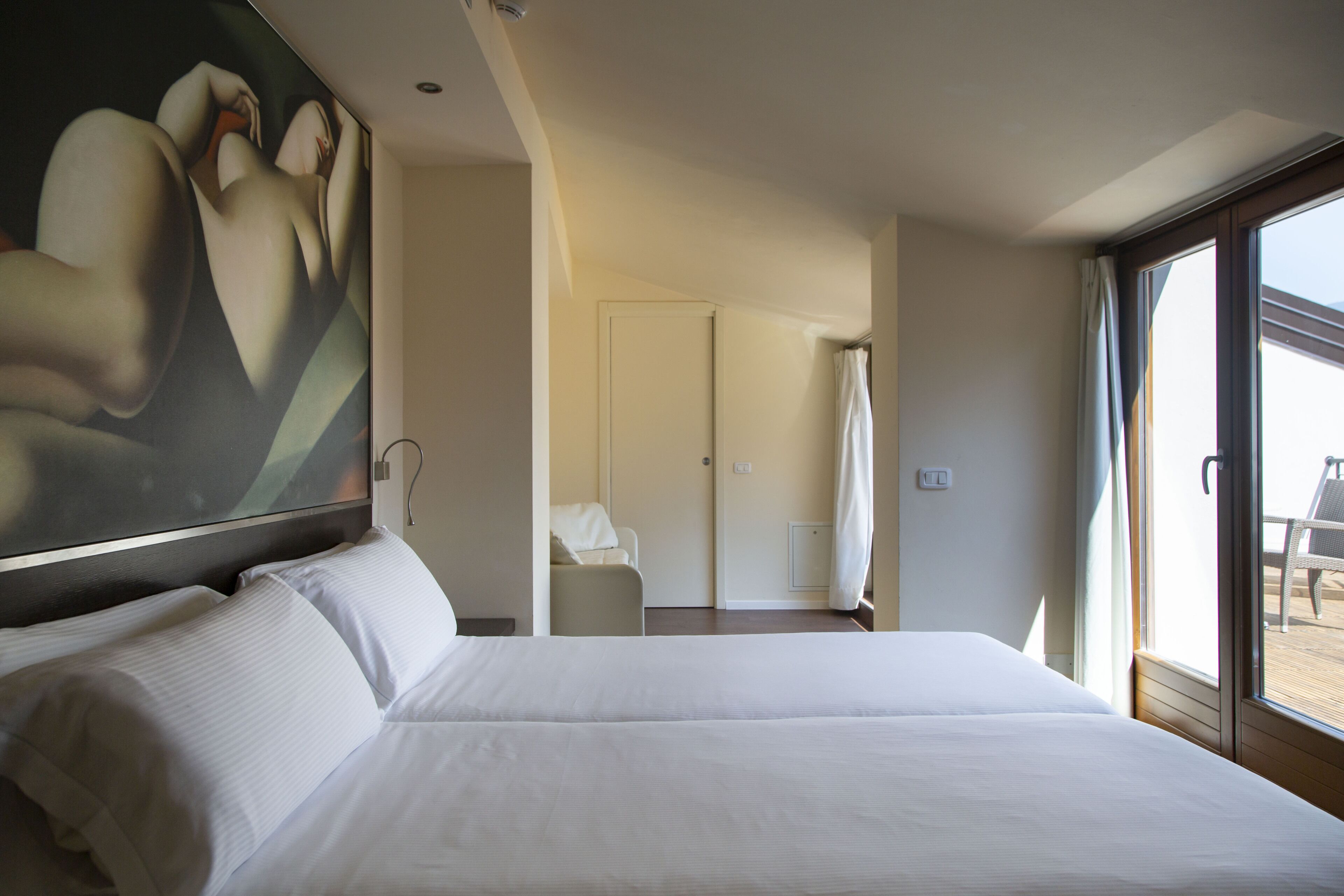 A hotel room with a large bed, a painting on the wall, a door leading to a balcony, and a chair.