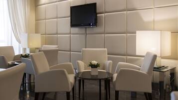 Ein modernes Hotelzimmer mit einem großen Flachbildfernseher, der an einer gepolsterten Wand befestigt ist, einem runden Glastisch mit vier Stühlen und zwei Nachttischlampen.