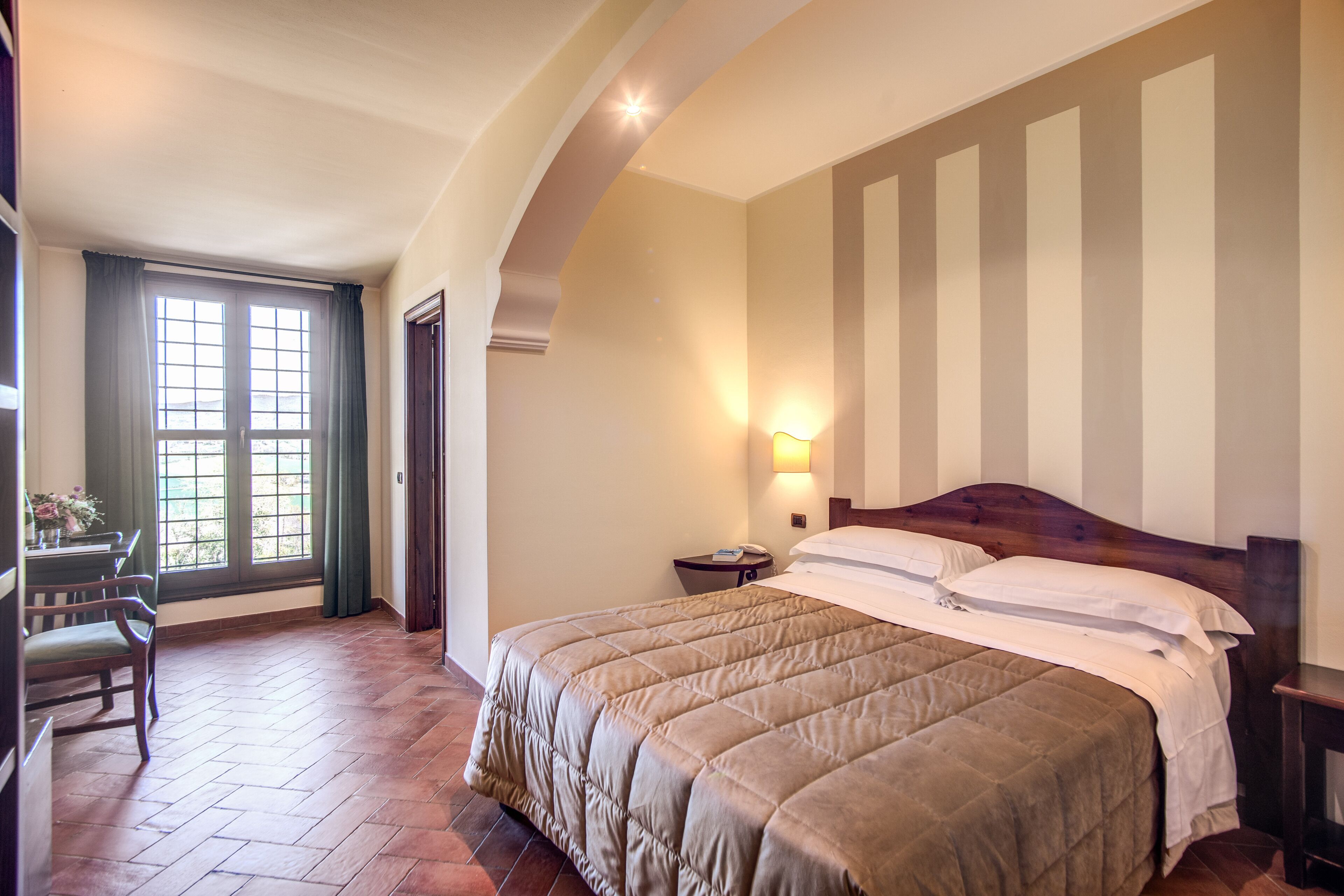 Habitación de hotel con una cama grande, un escritorio con silla, una ventana con cortinas y una lámpara fijada en la pared.