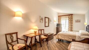 Une chambre d’hôtel avec un plancher en bois, un lit recouvert d’une couette beige, un bureau avec une lampe, deux chaises et un téléviseur.
