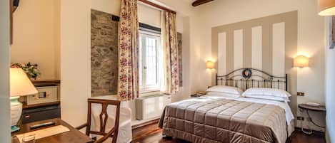 Un dormitorio con cama, escritorio y silla, lámpara, ventana con cortinas y pared de piedra.