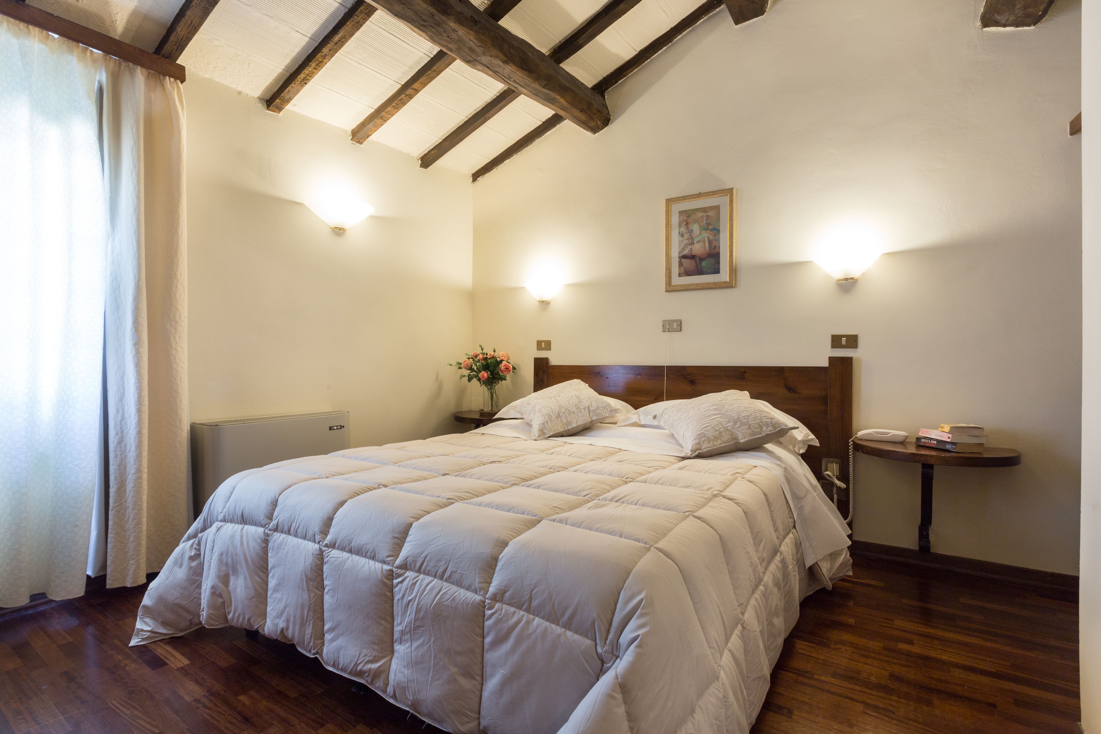 A bedroom with a large bed, wooden beams on the ceiling, a bedside table with a lamp, and a small round table with books and a phone.