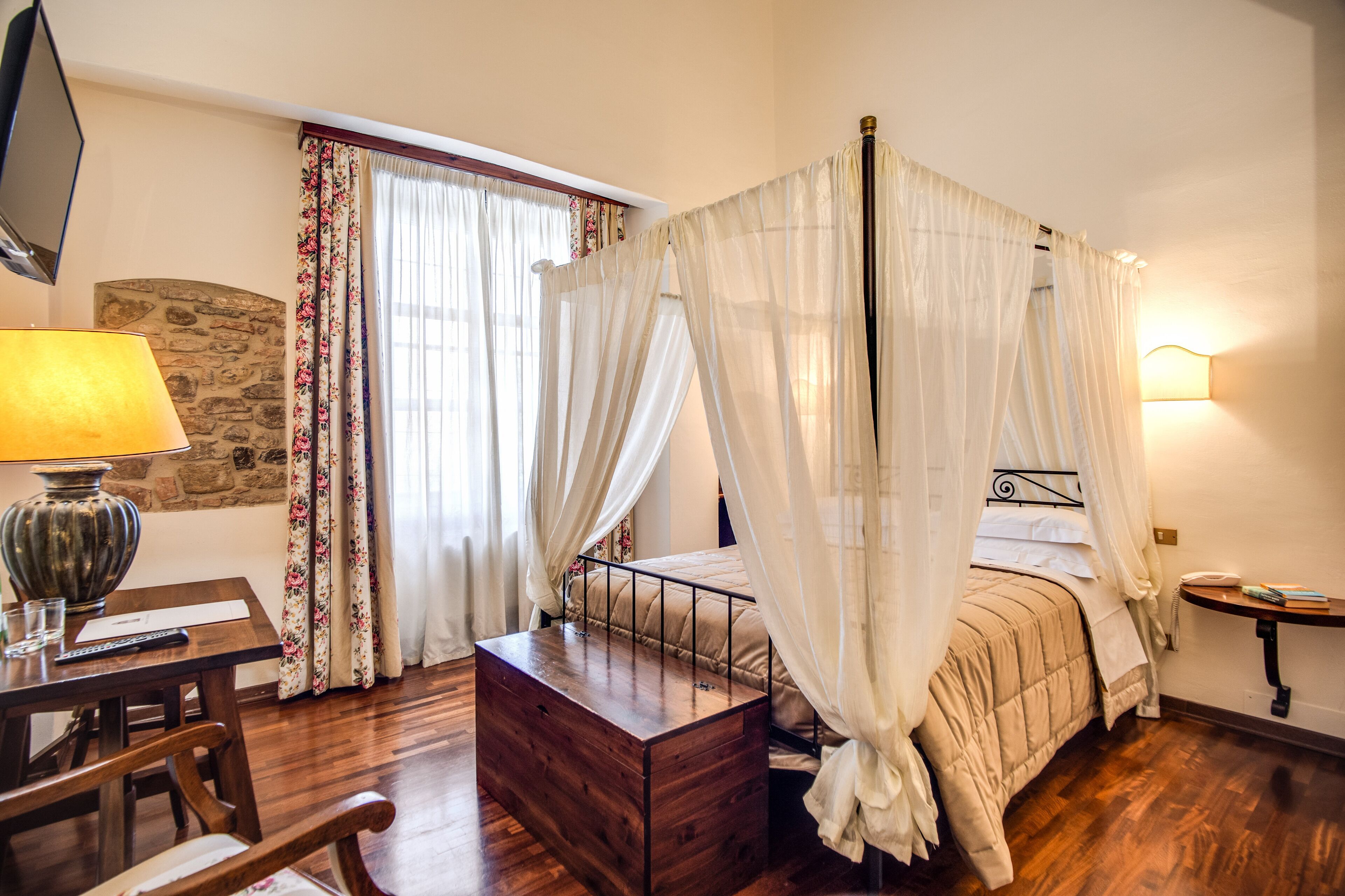 A four-poster bed with a canopy, a wooden bedside table, a lamp, a stone wall, and a window with curtains.