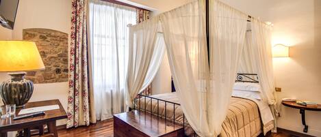 A four-poster bed with a canopy, a wooden bedside table, a lamp, a stone wall, and a window with curtains.