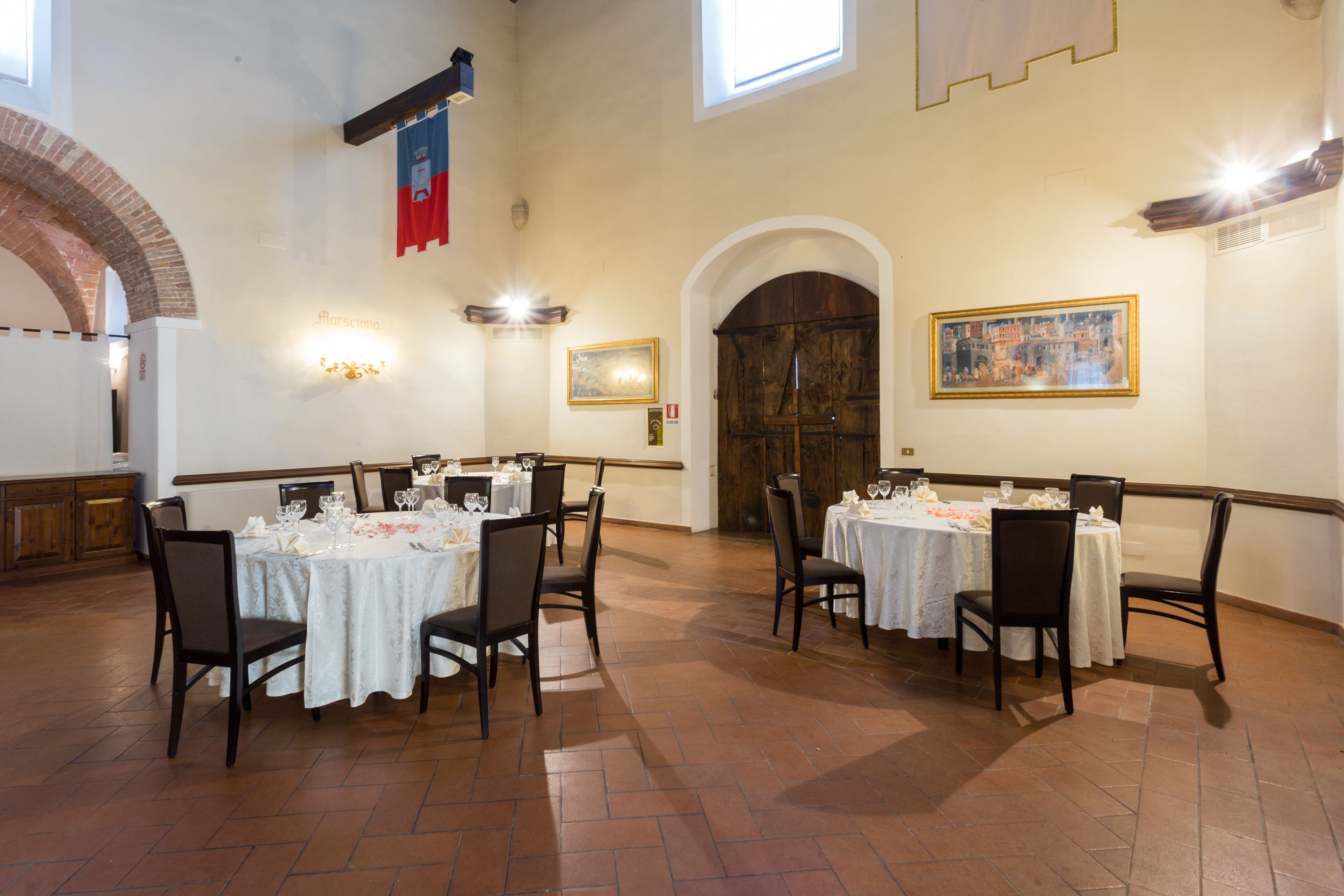 A dining area with tables set for four, each with white tablecloths and dark chairs. There are two windows providing natural light, and a large wooden door with a painting above it.