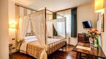 A four-poster bed with a canopy, a bedside table with a lamp, a flat-screen TV, and a vase with flowers.