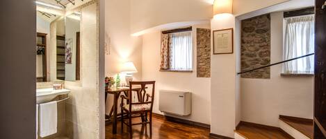 A bathroom with a shower, a sink, and a mirror. There is a wooden chair and a table with a lamp. The room has wooden flooring and a staircase with a stone wall.