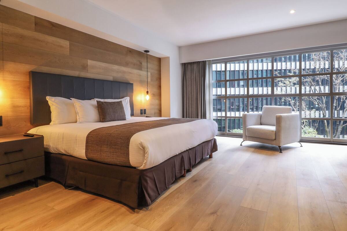 A modern bedroom with a large bed, a nightstand, a chair, and a large window with a view of buildings.